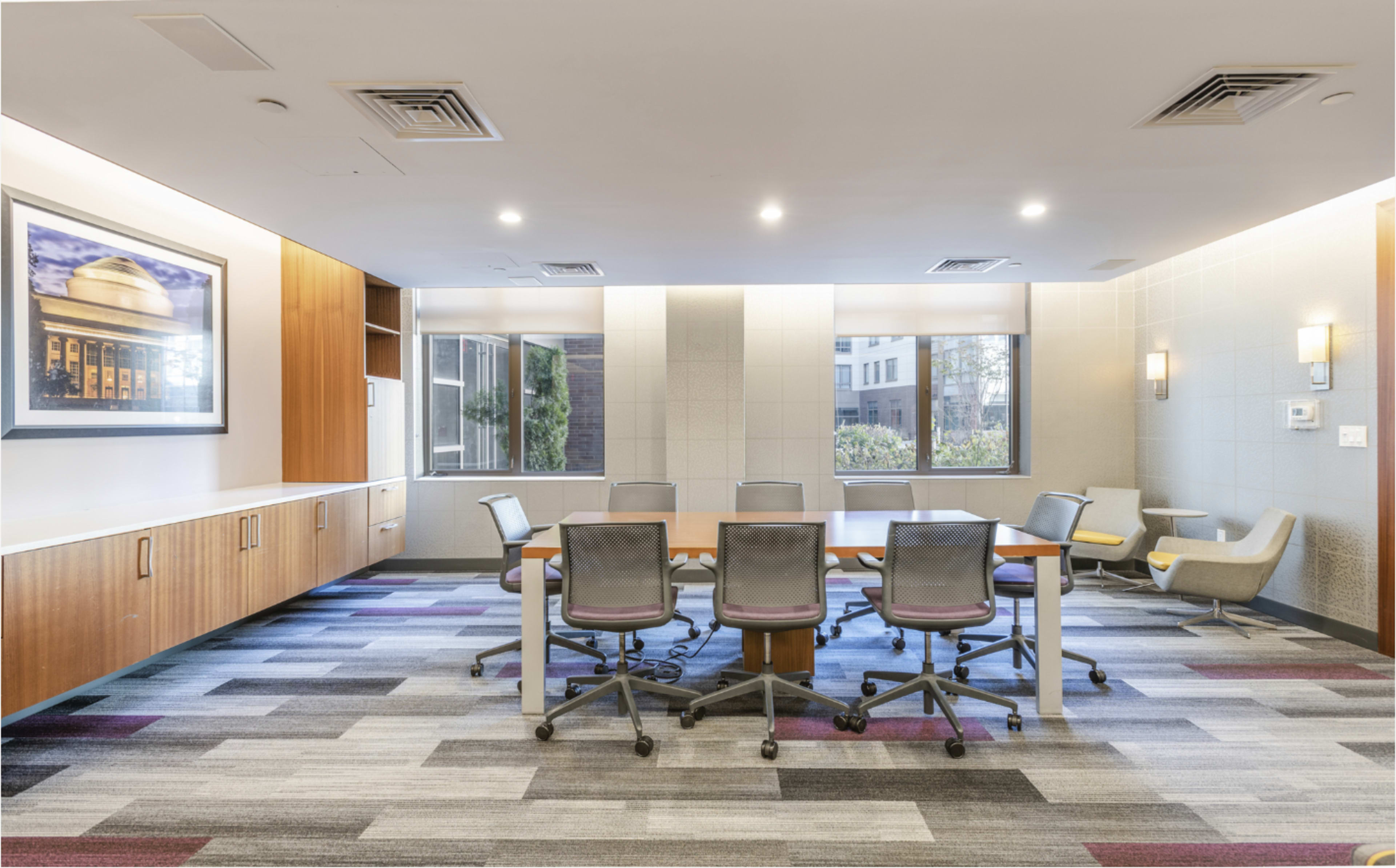 The image shows a modern conference room with a round table surrounded by chairs, large windows providing natural light, and a wall featuring wooden cabinets and artwork.