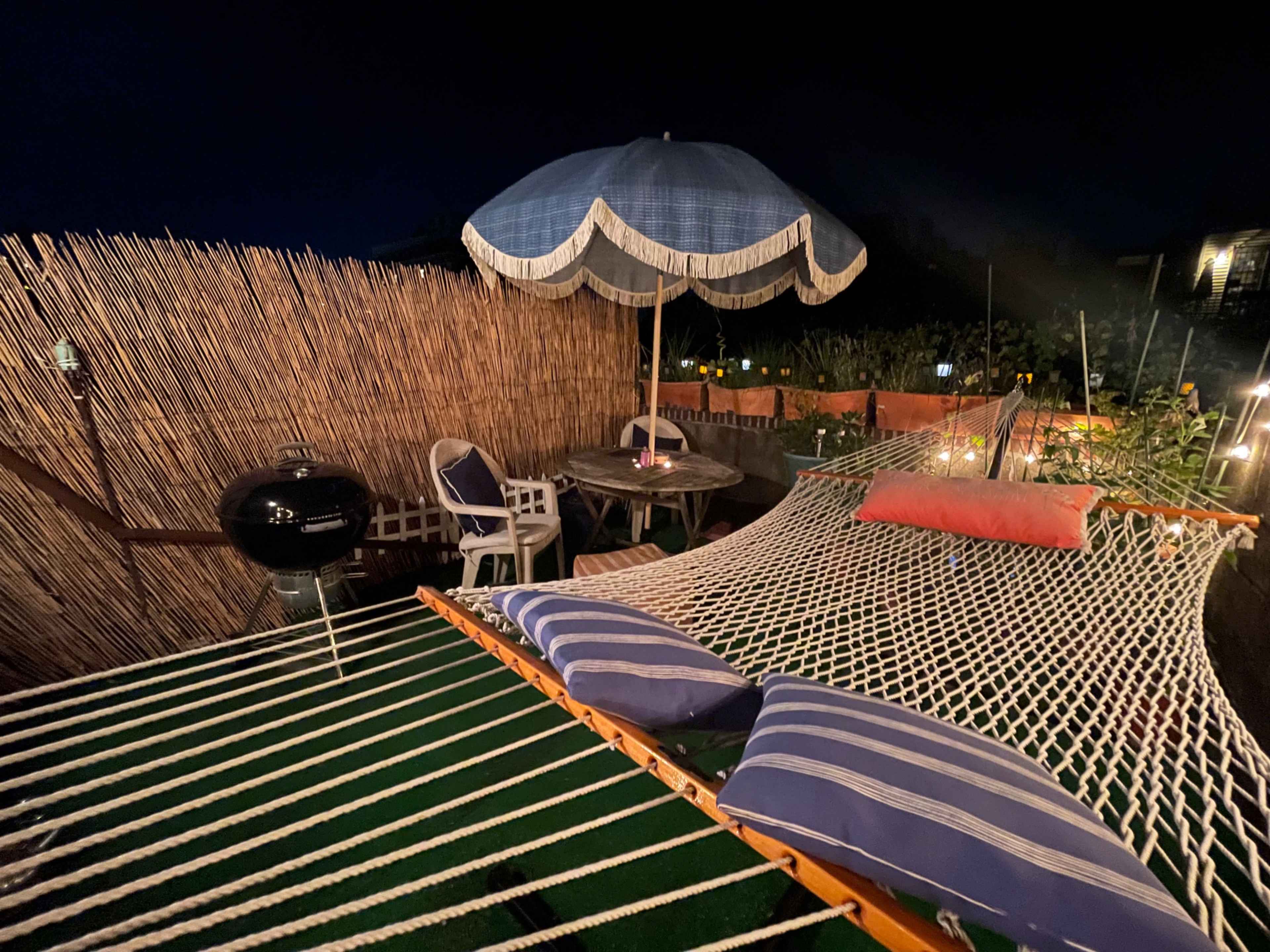 A hammock with striped cushions is set up under a blue umbrella in a dimly lit outdoor space surrounded by a bamboo fence and a small table with chairs.