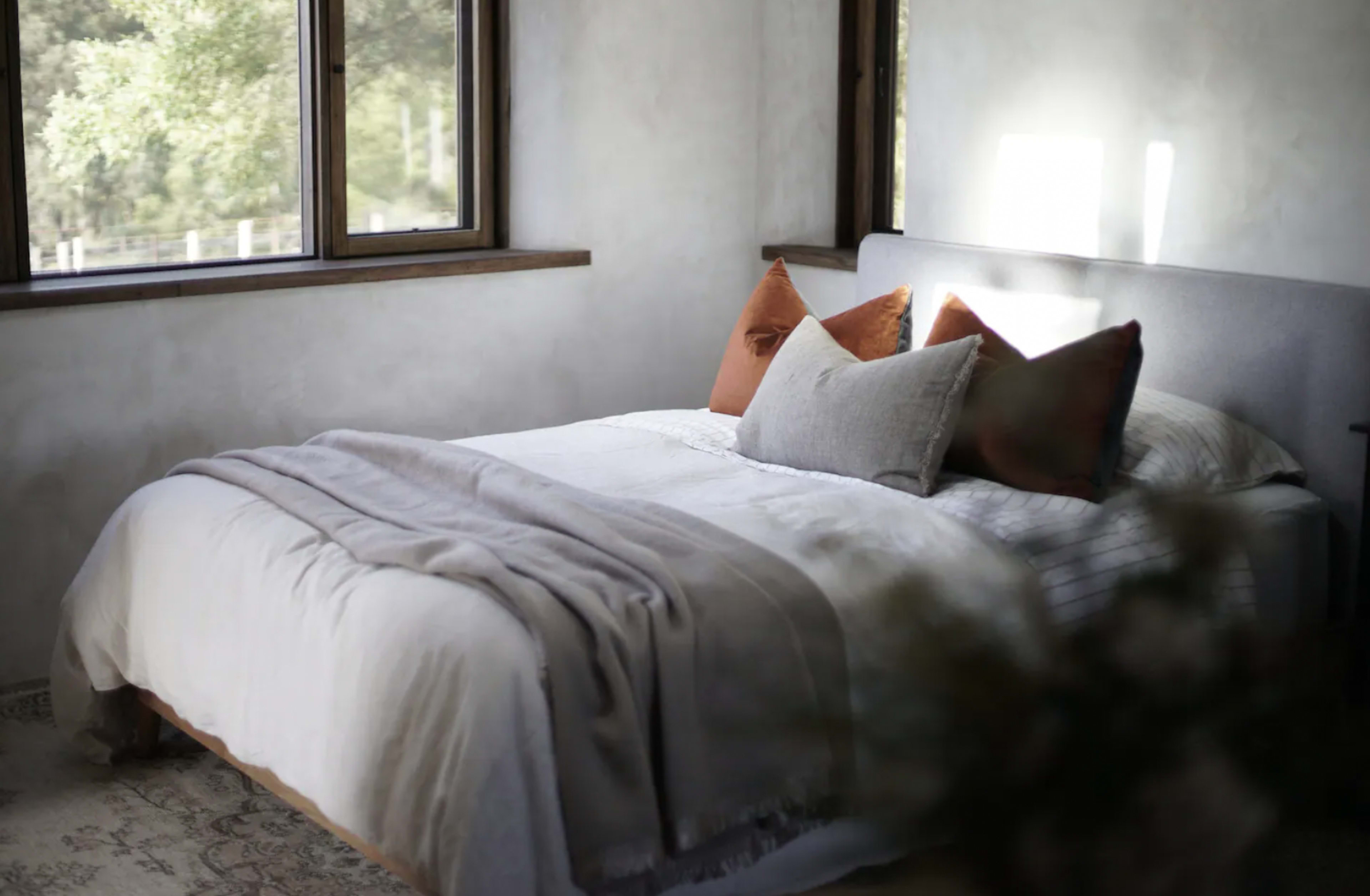 A neatly made bed with decorative pillows placed against a light-colored wall and large windows letting in natural light.
