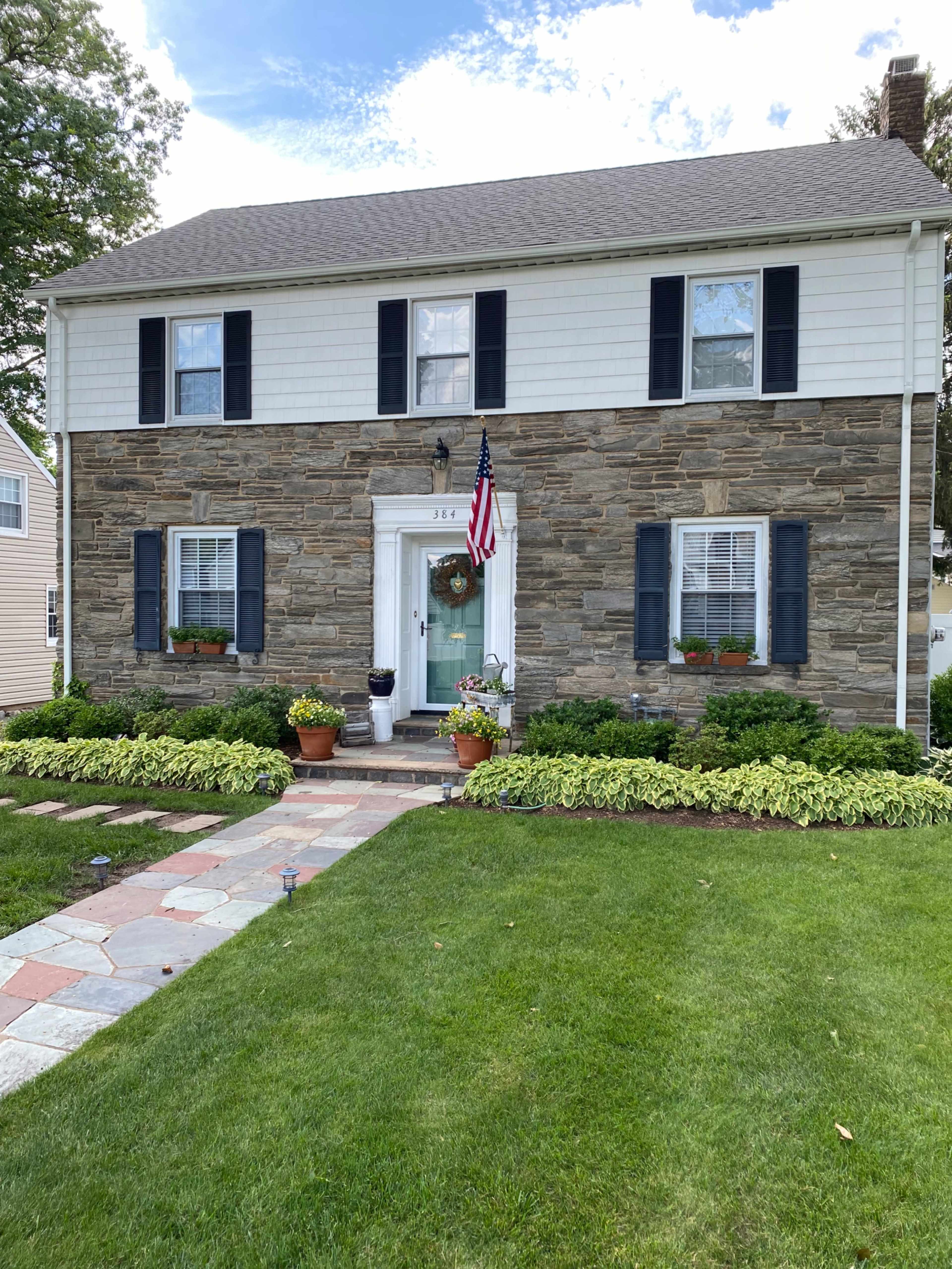 A two-story house with a stone facade, white upper siding, and a neatly landscaped front yard featuring potted plants and a walkway made of stone slabs.