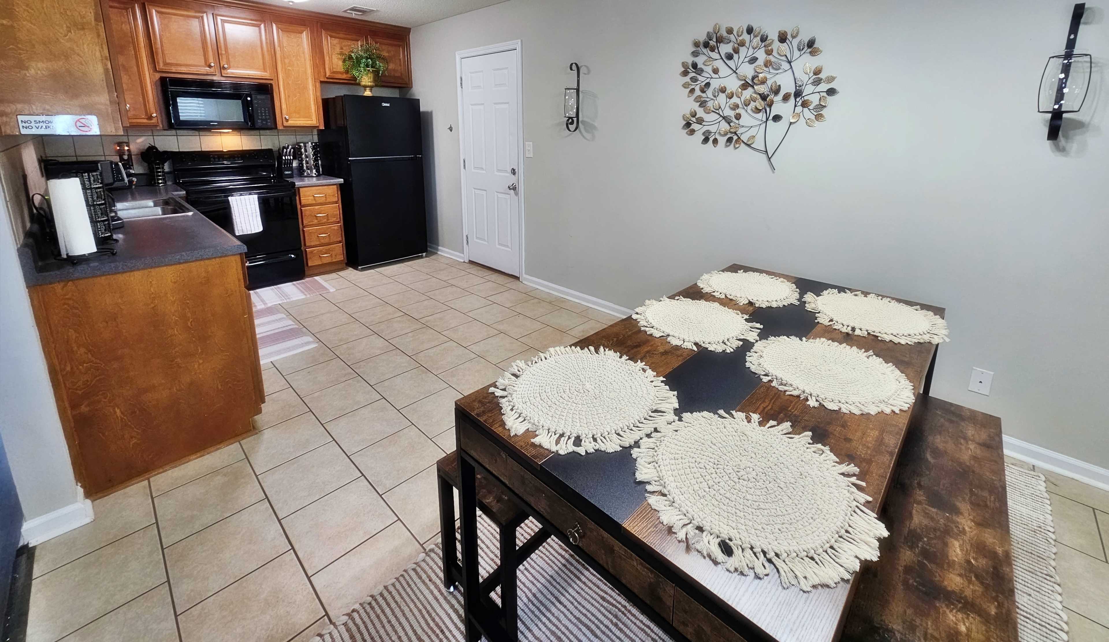 The image shows a kitchen and dining area featuring wooden cabinets, a black refrigerator, and a dining table set with woven placemats.