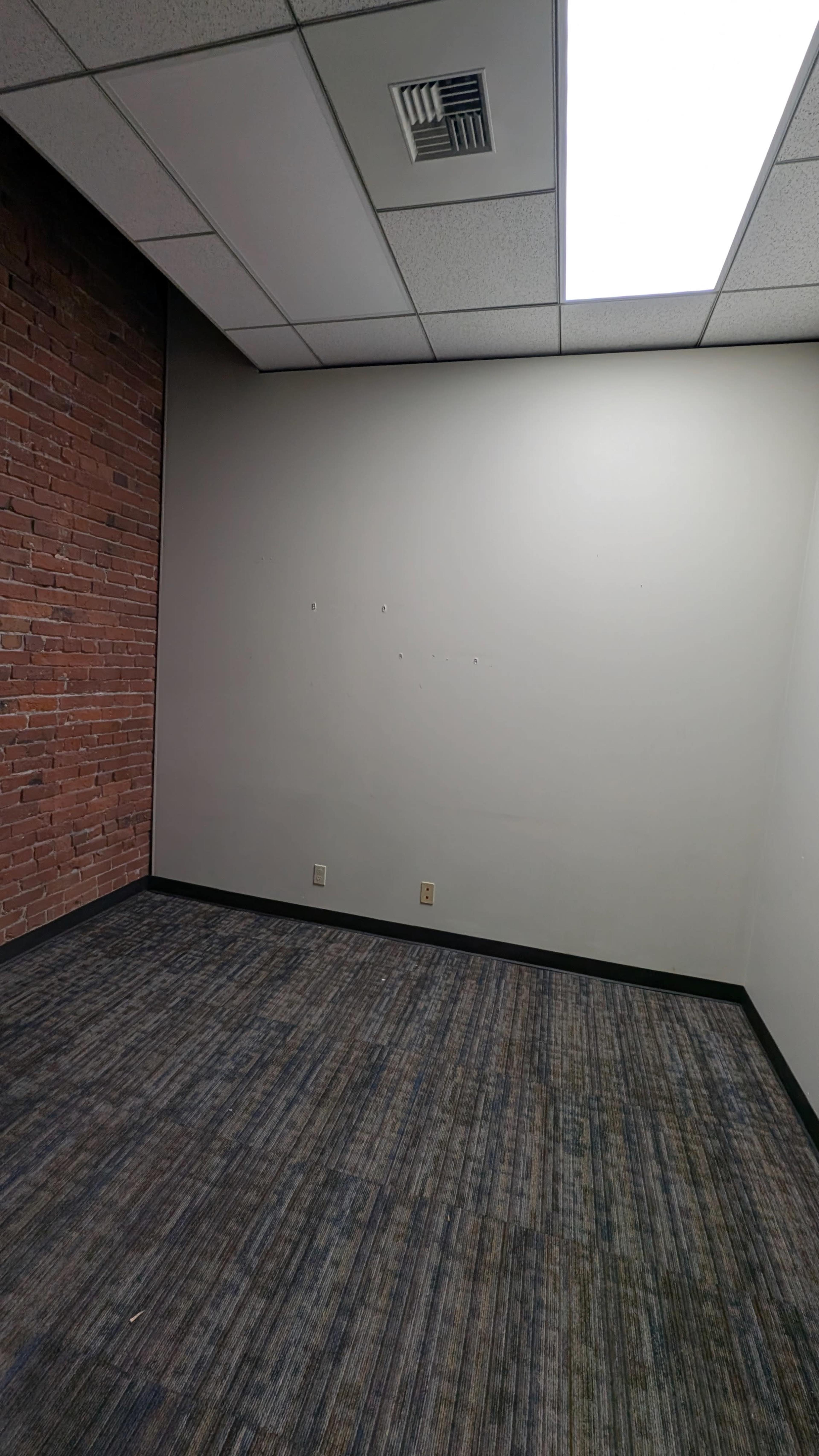 The image shows a vacant office space with one brick wall and two empty walls, featuring a carpeted floor and fluorescent lighting above.