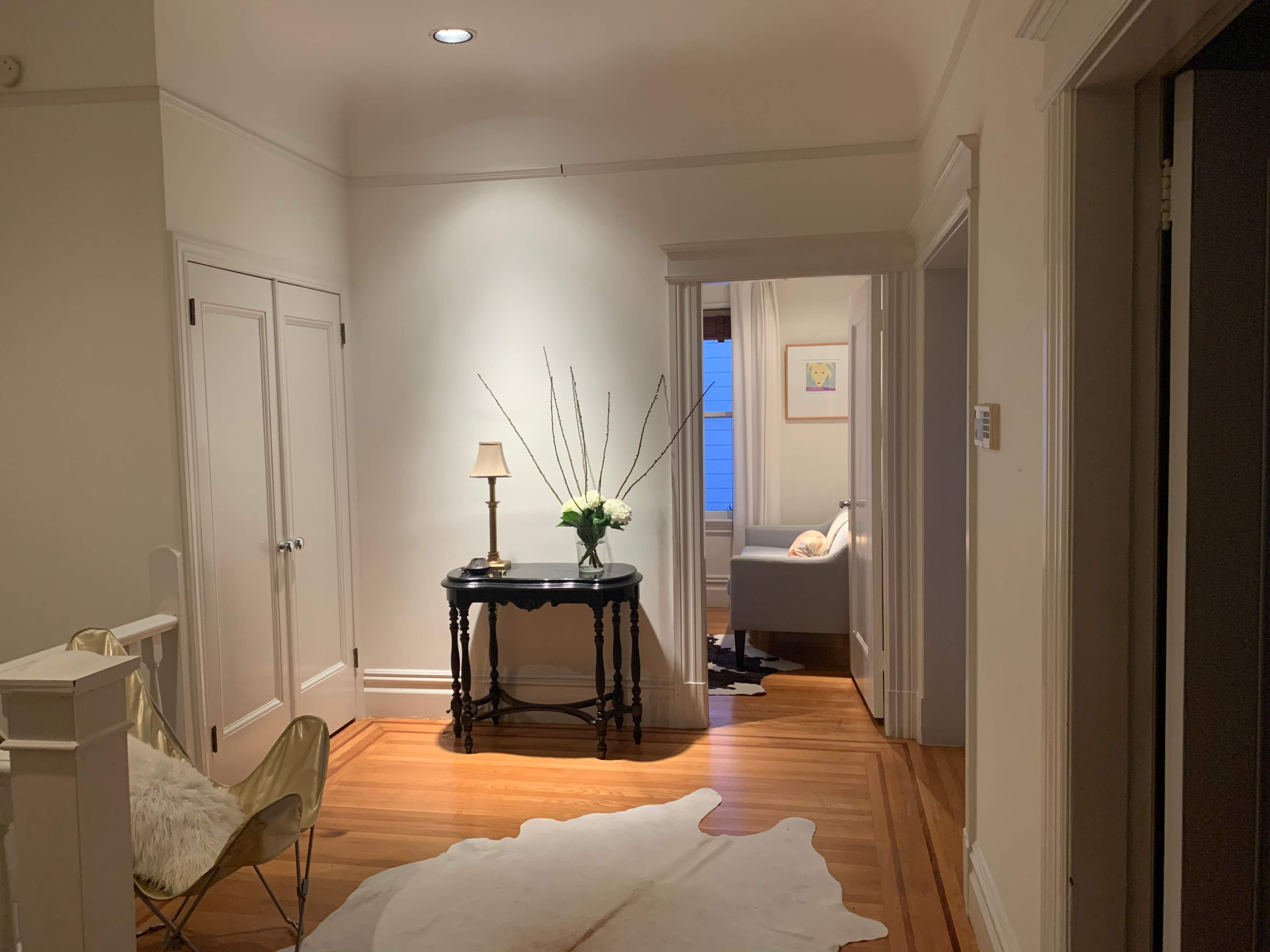 A well-lit hallway with a table and a lamp, leading to a room with a couch and a window.