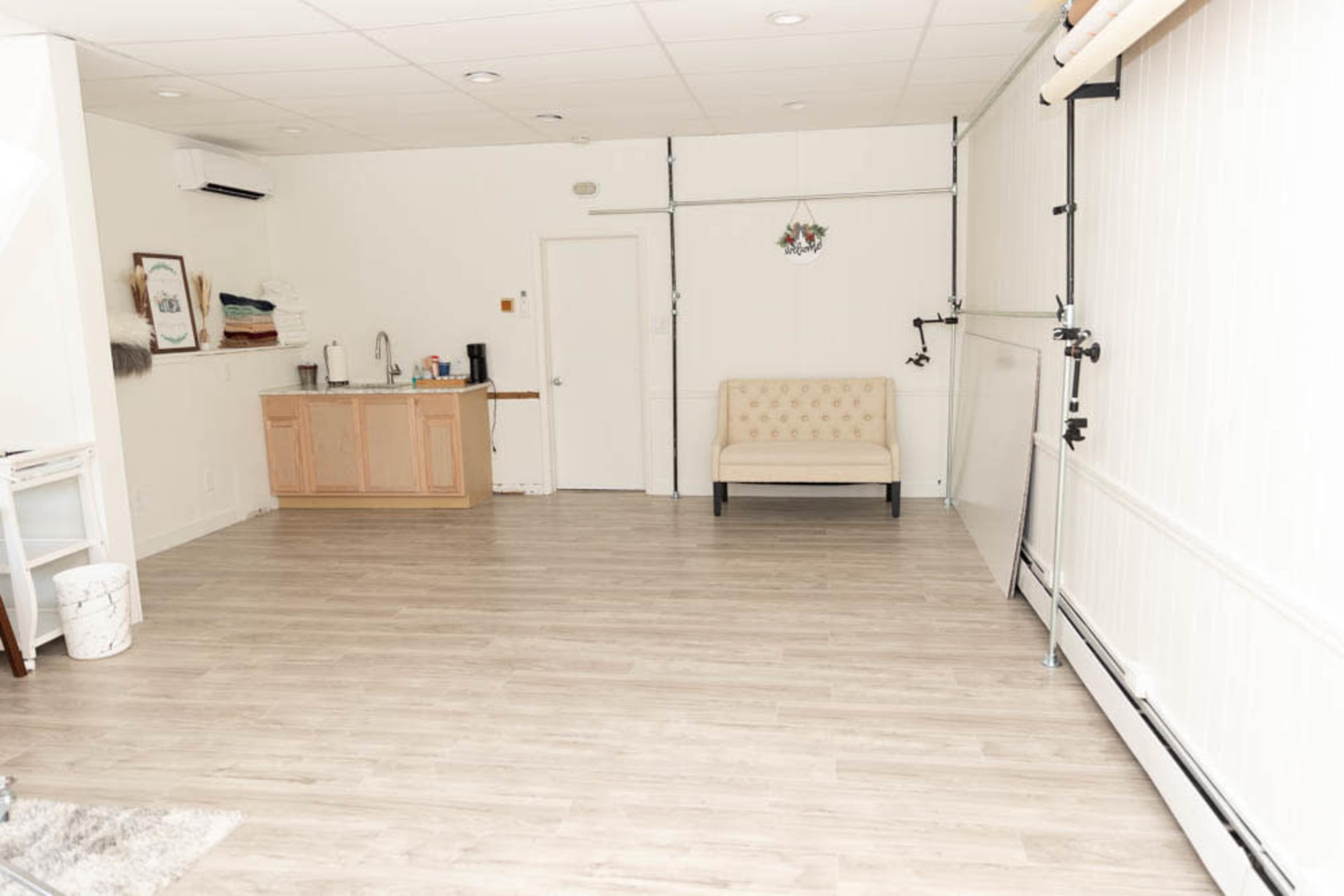 A spacious room with light wood flooring, a kitchenette in the back, a decorative couch, and a wardrobe rack along one wall.