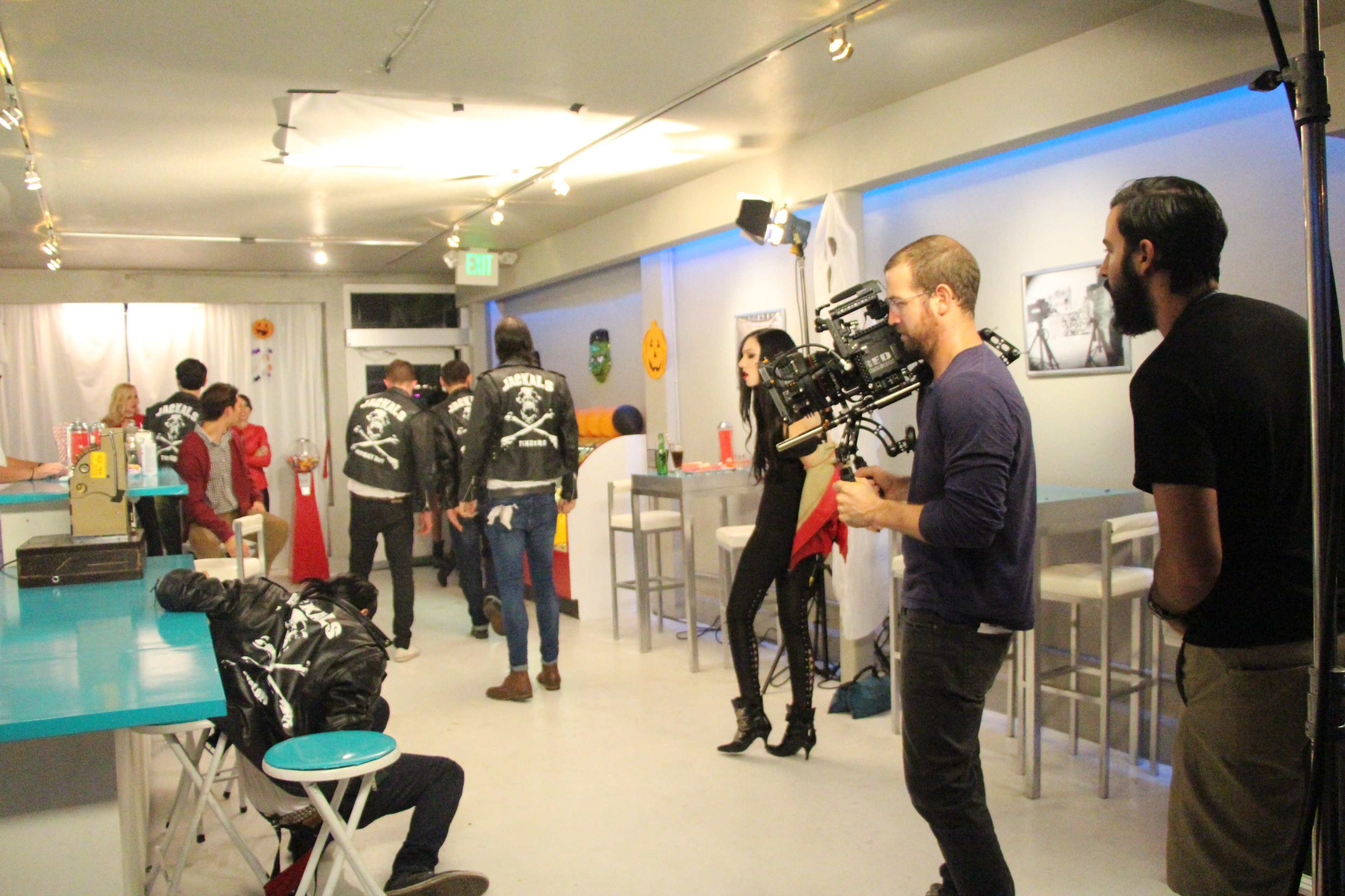 A film crew captures a scene in a brightly lit room featuring actors in leather jackets while another actor is seated in front, surrounded by various props and decorations.