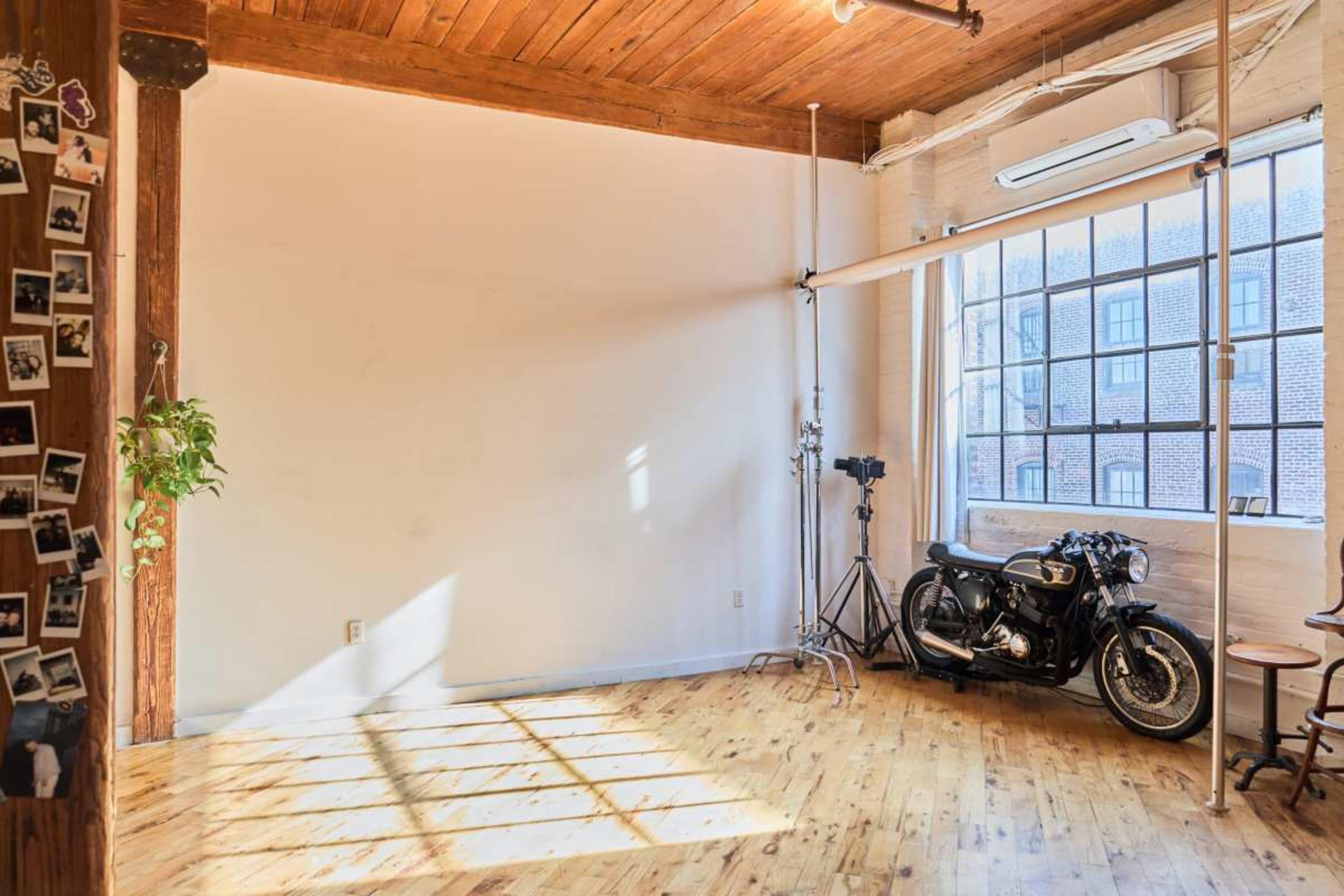 The image shows a spacious, brightly lit room with wooden flooring, large windows, and a vintage motorcycle parked against a wall.