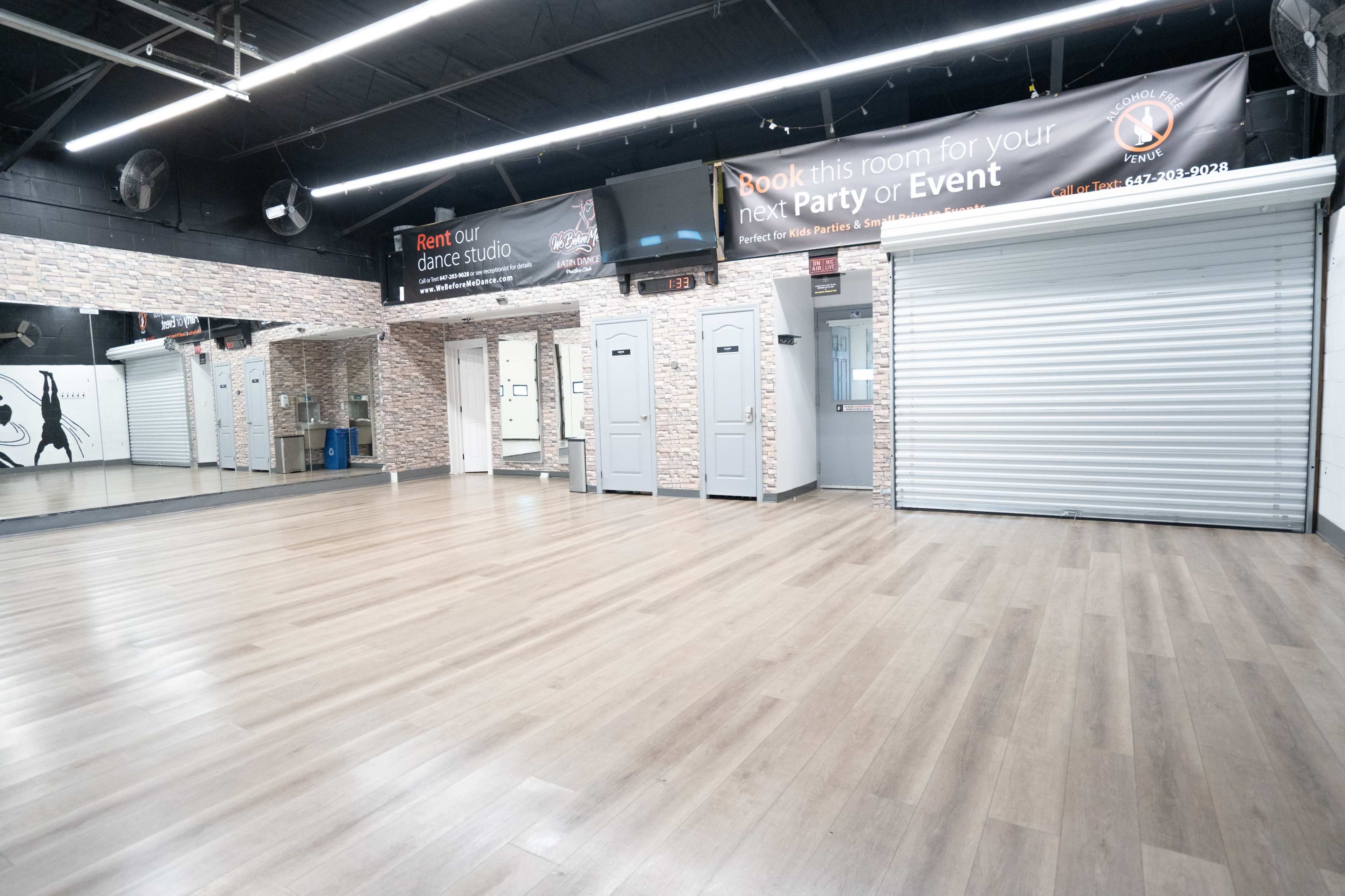 The image shows an empty dance studio with wooden flooring, mirrored walls, and a roller shutter door, along with promotional banners on the walls.