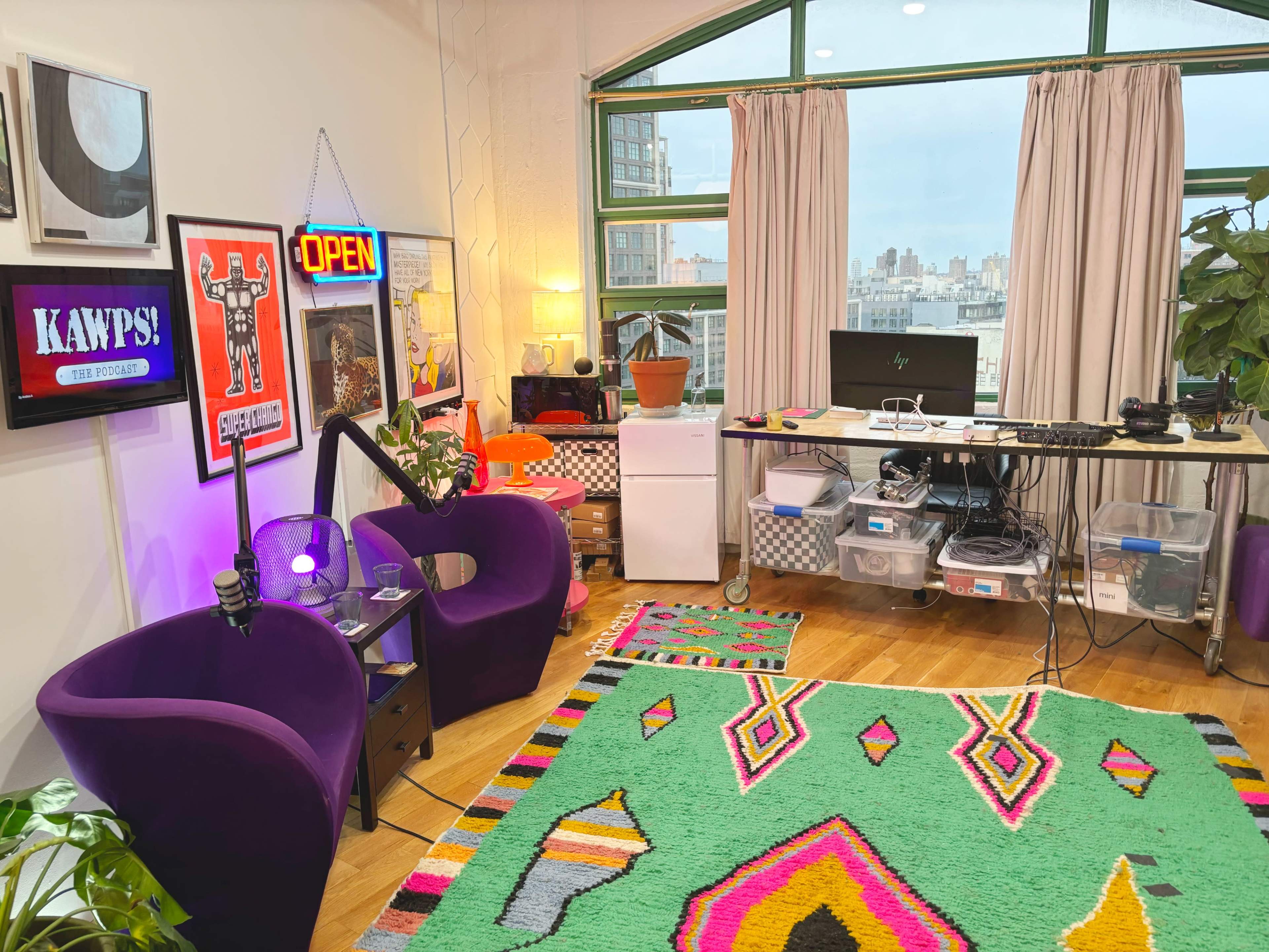 The image shows a modern workspace with purple chairs, a colorful rug, a desk with a computer and storage bins, and large windows overlooking a city skyline.