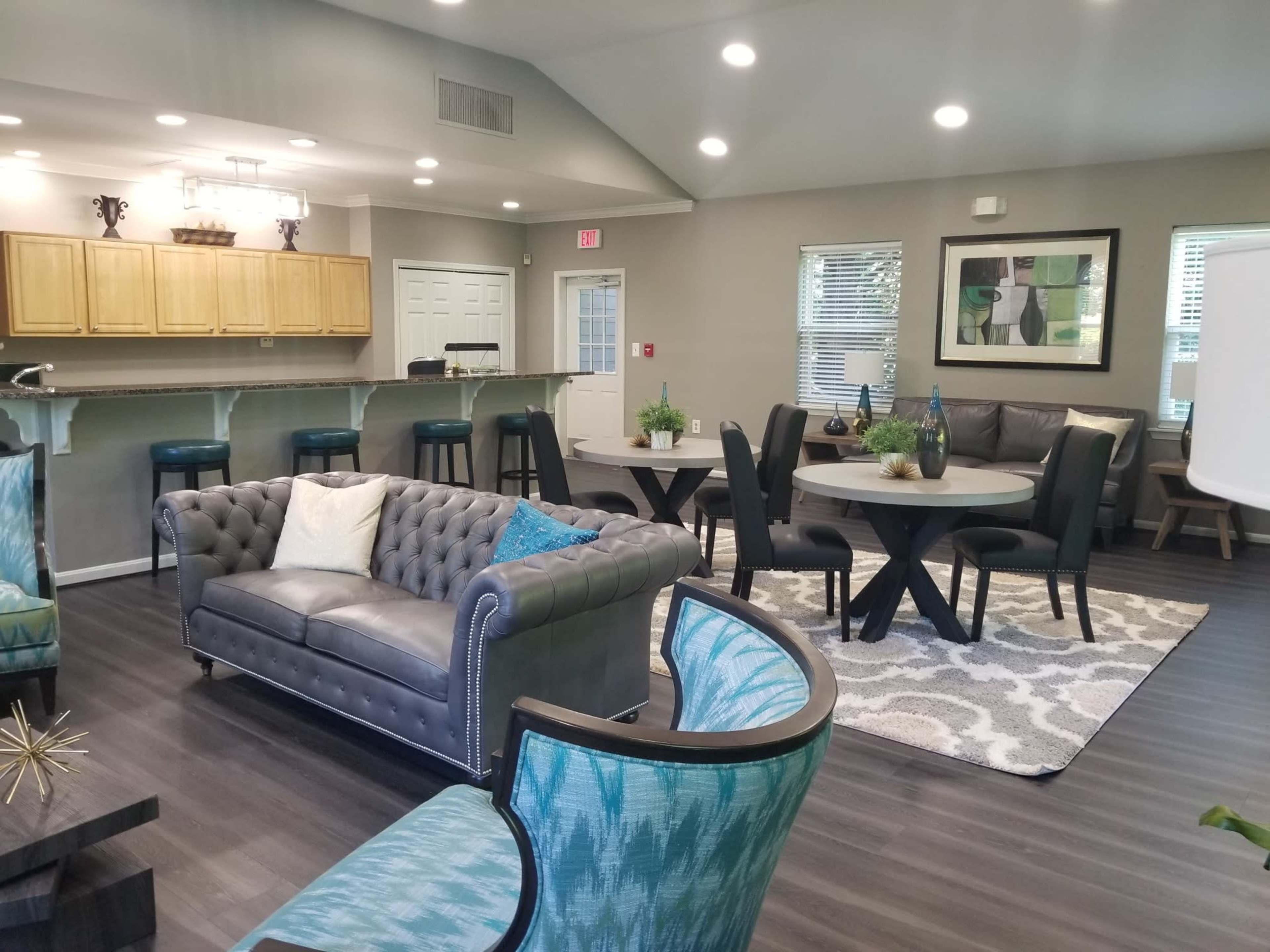 The image shows a modern lounge area with a gray sectional sofa, two round dining tables, and a kitchen bar with stools, all arranged around a plush area rug.