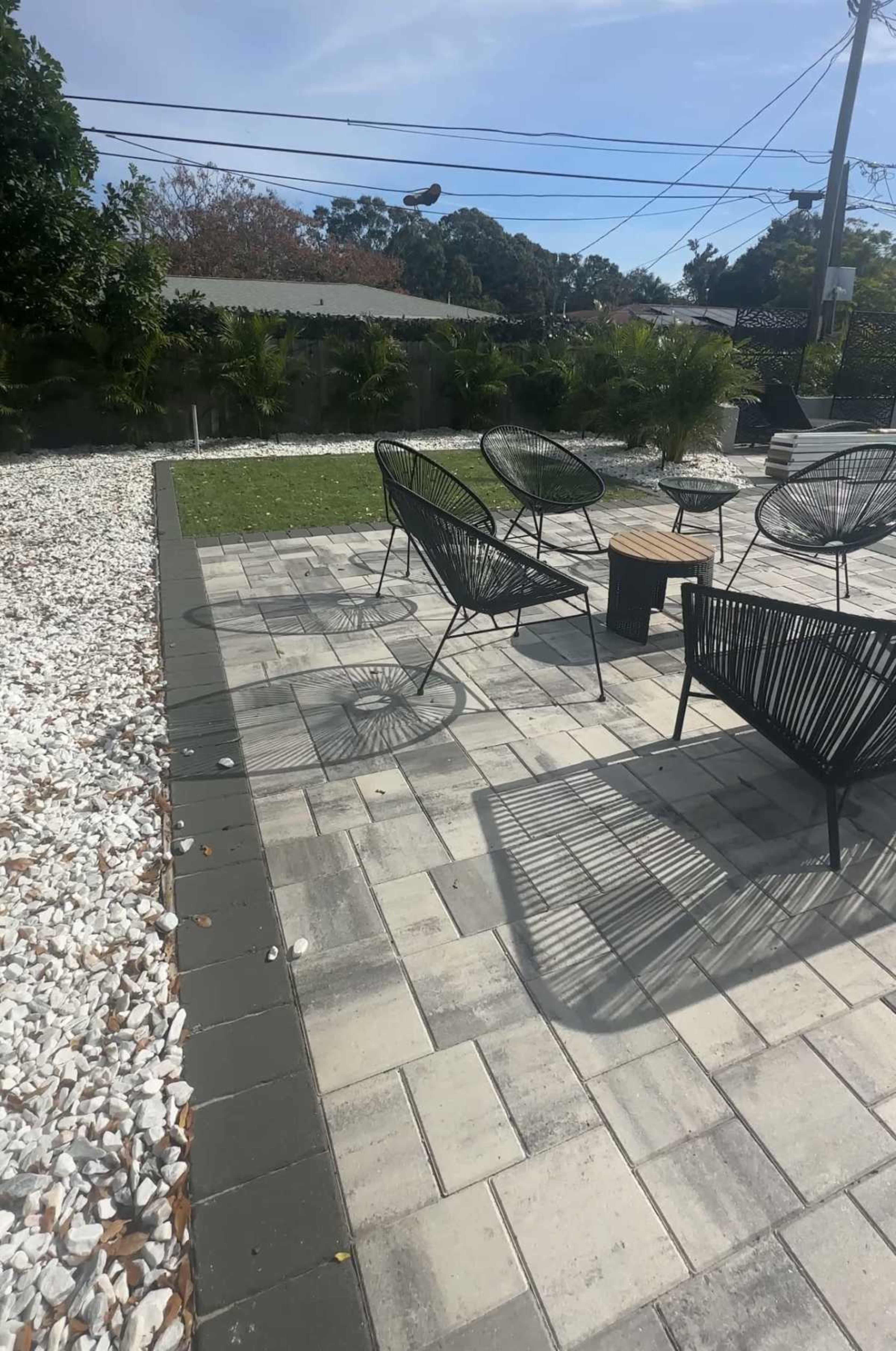 The image shows a patio area paved with square stones, featuring several black wire chairs and a small wooden table, surrounded by greenery and white gravel.