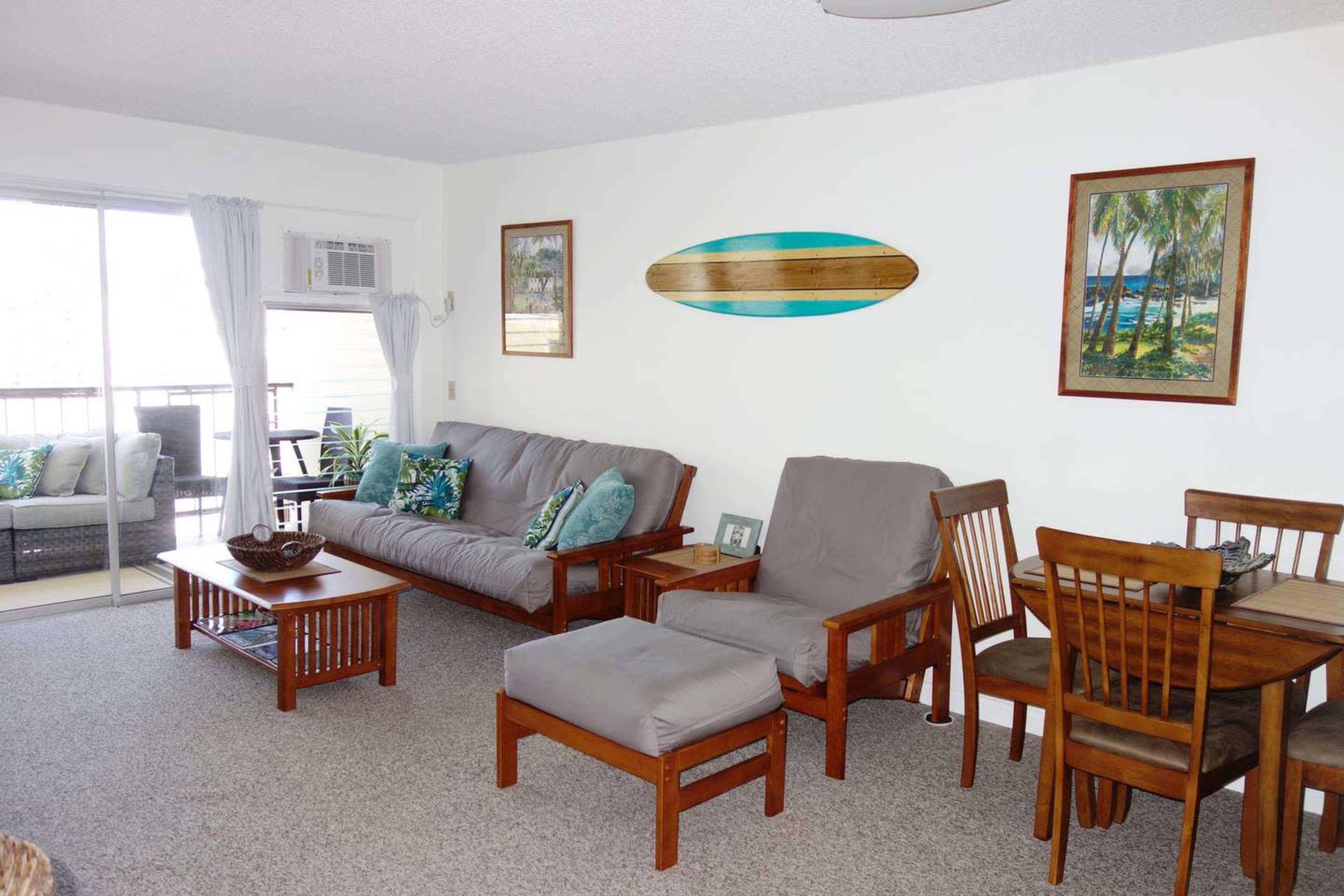 A living room with a gray futon, wooden furniture, and tropical-themed artwork, adjacent to a balcony area with sliding glass doors.