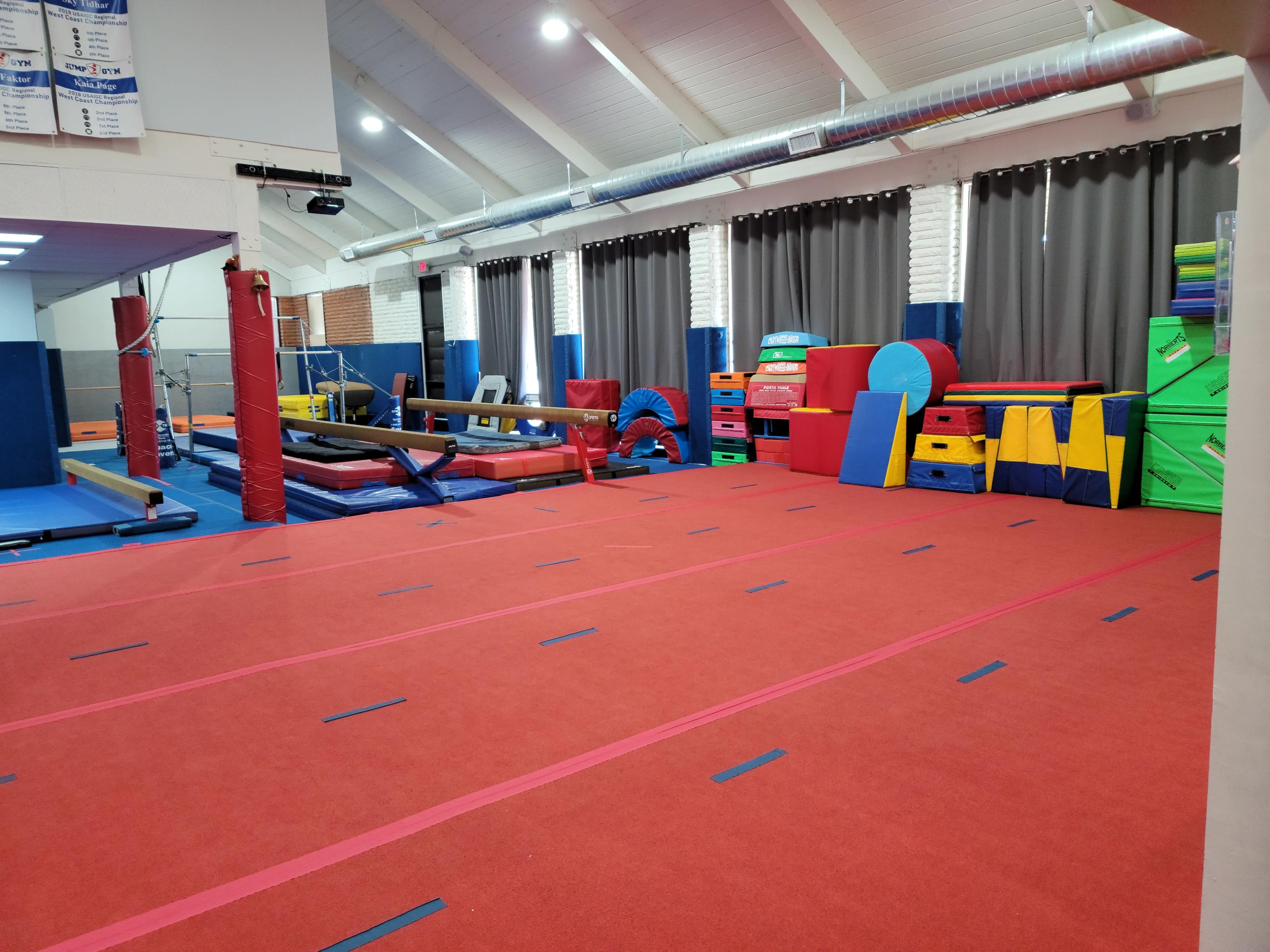 A gymnastics gym with red carpet flooring, various training equipment, and colorful mats arranged along the walls.