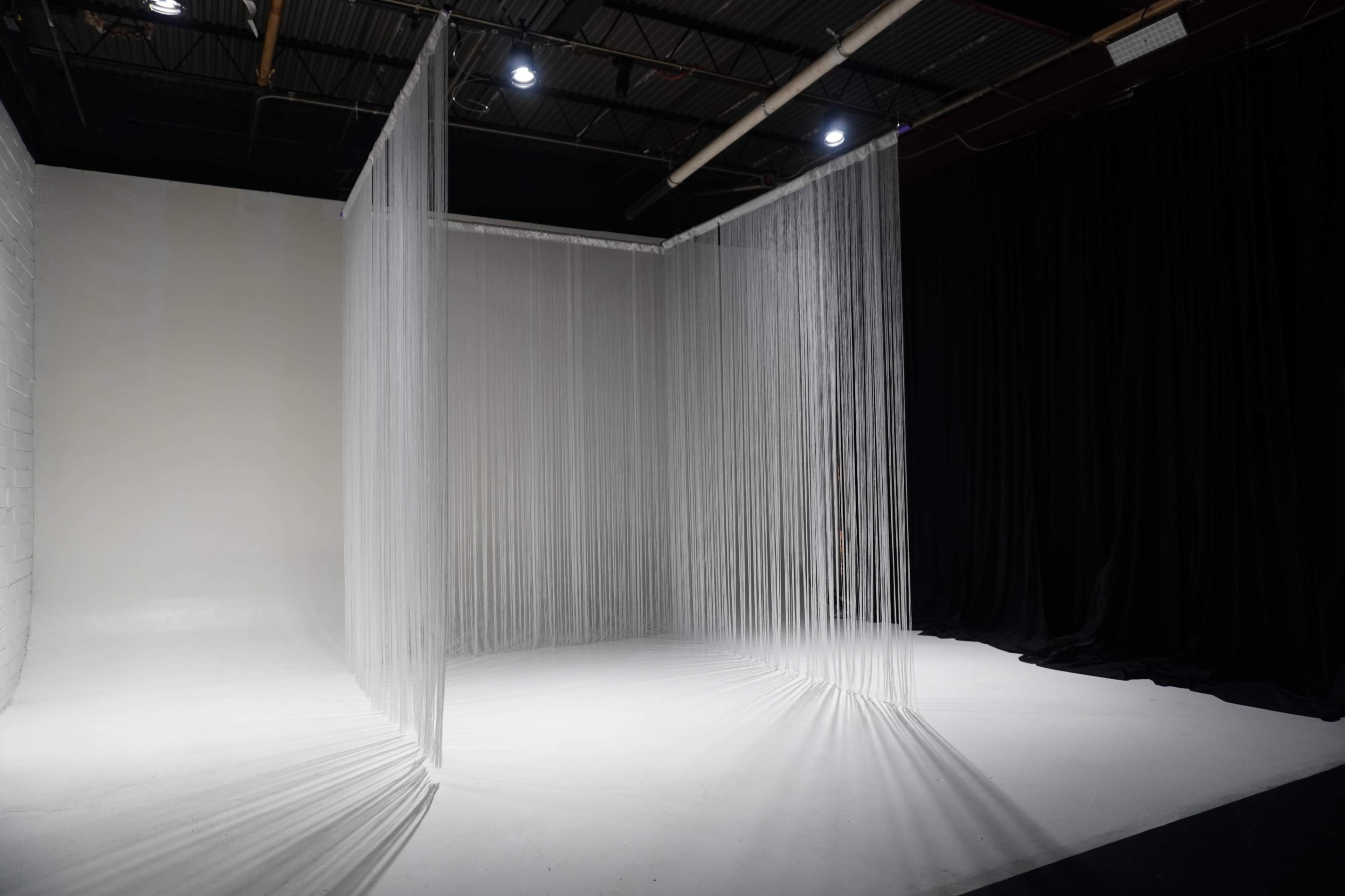 The image shows a minimalist studio space with sheer, vertical curtains dividing the area against a backdrop of white and black walls.