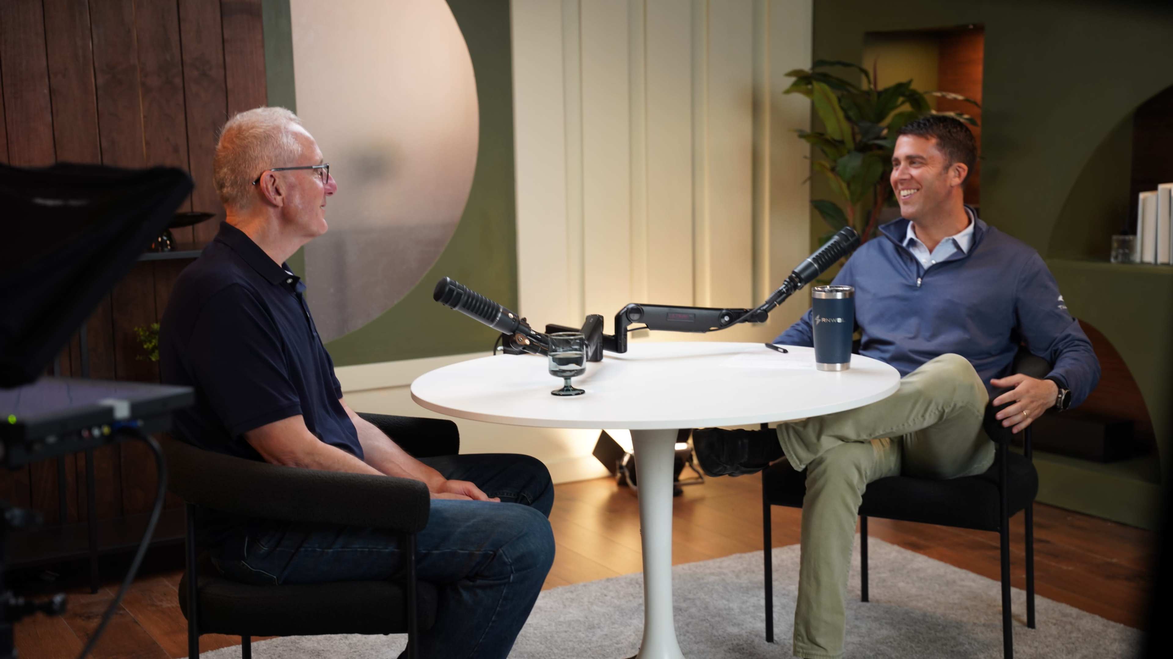 Two men are seated at a round table with microphones, engaged in conversation in a modern indoor setting.