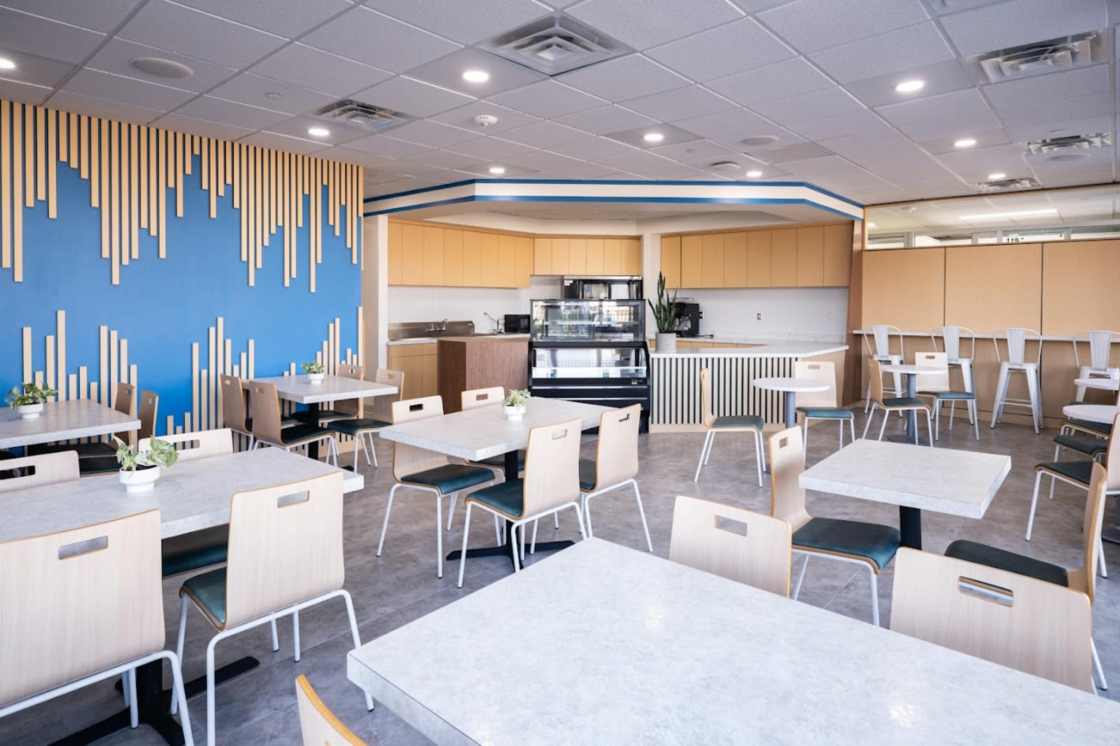 The image shows a modern cafeteria with tables and chairs arranged neatly, and a service counter visible in the background.