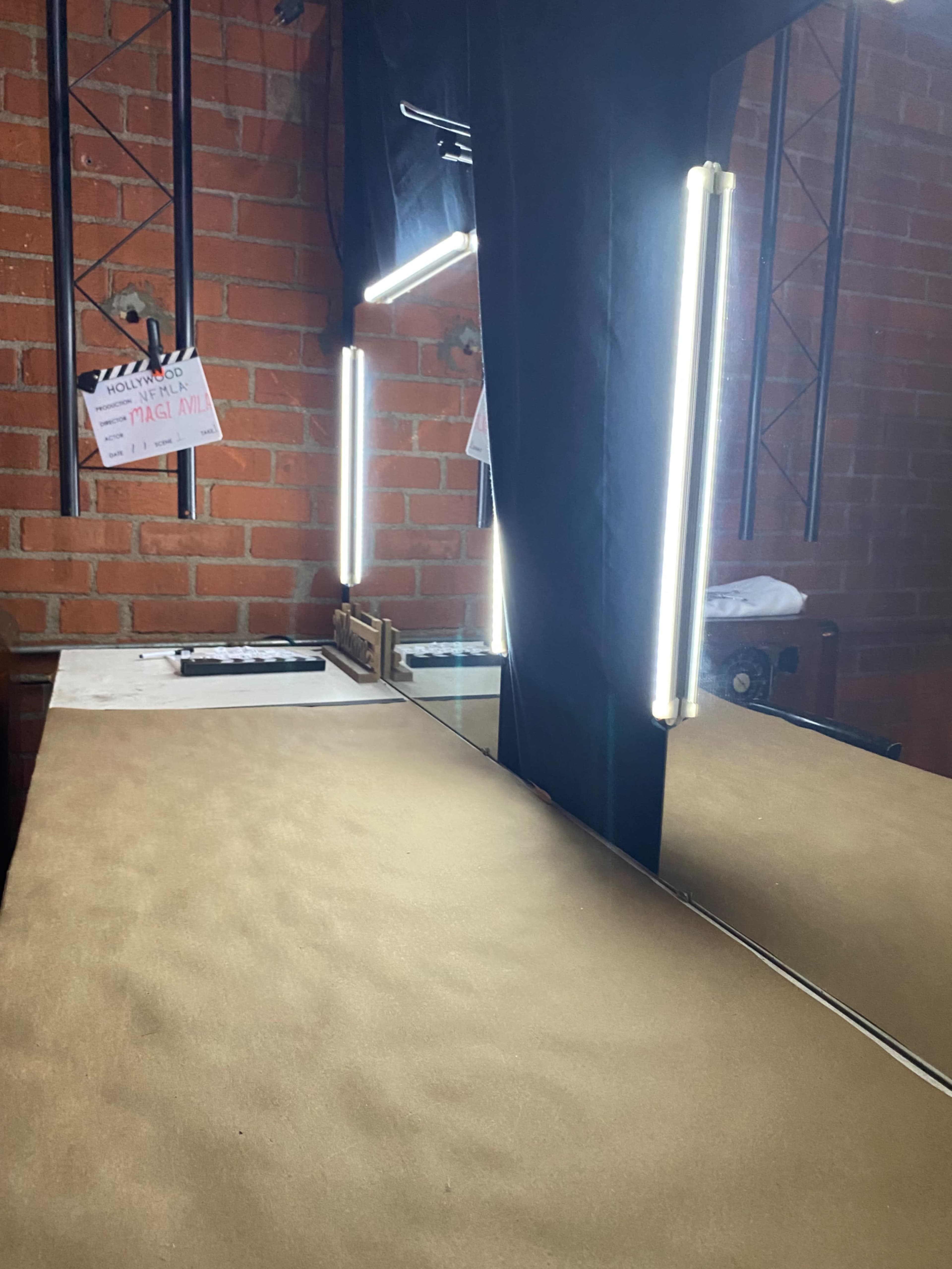 A vanity table with a large mirror surrounded by bright lights, set against a brick wall.