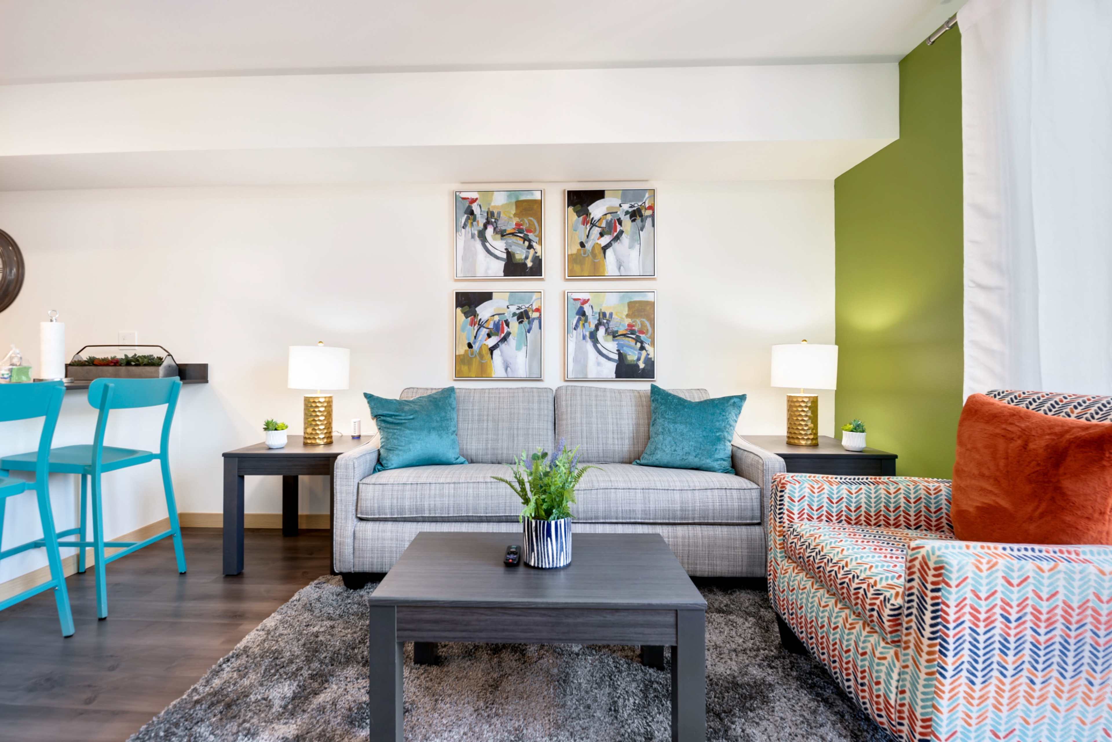 A modern living room with a gray sofa, an orange accent chair, and a coffee table, surrounded by colorful wall art and two lamps.