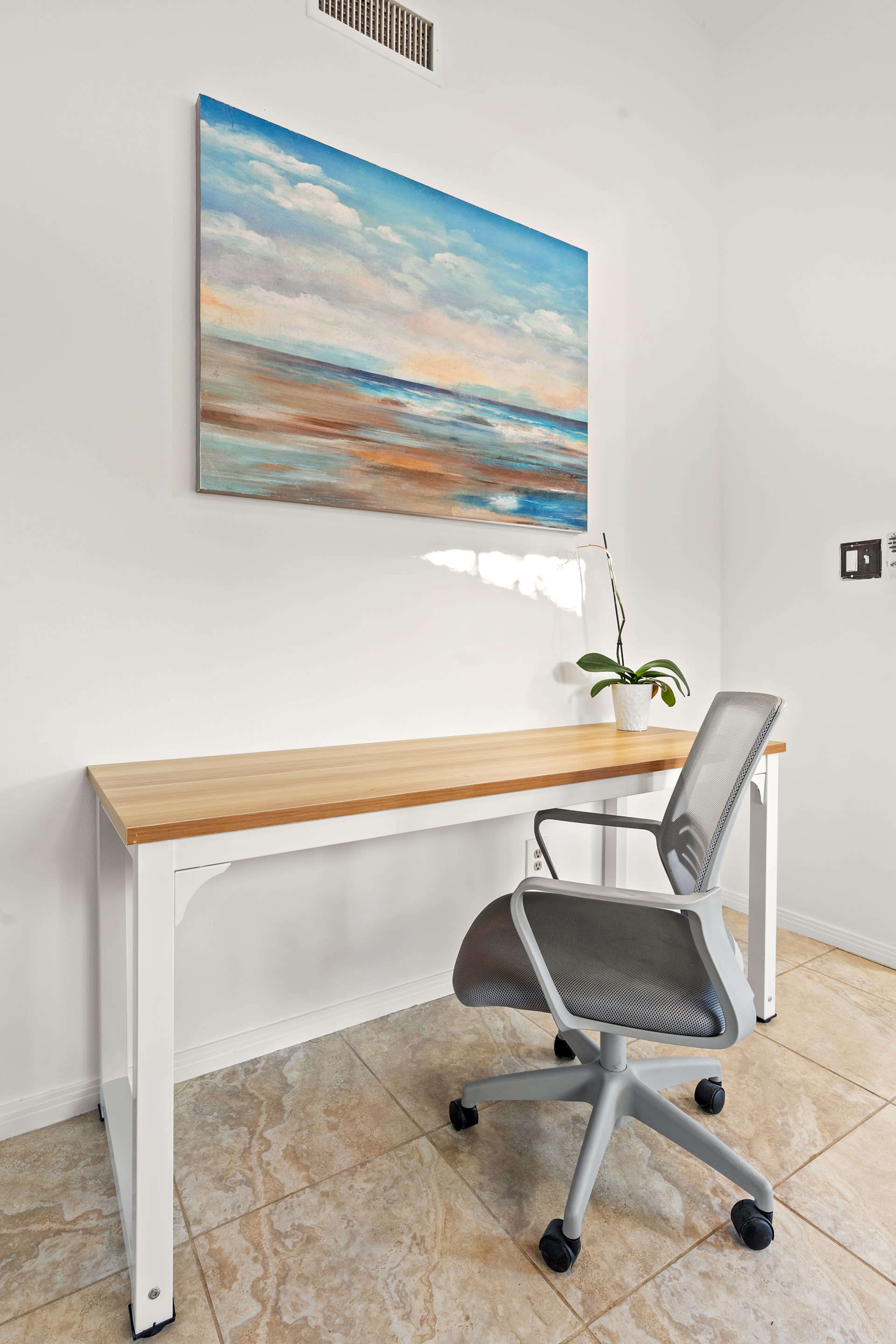 A minimalist workspace features a light wooden desk, an ergonomic chair, and a painting of a serene ocean landscape on the wall.