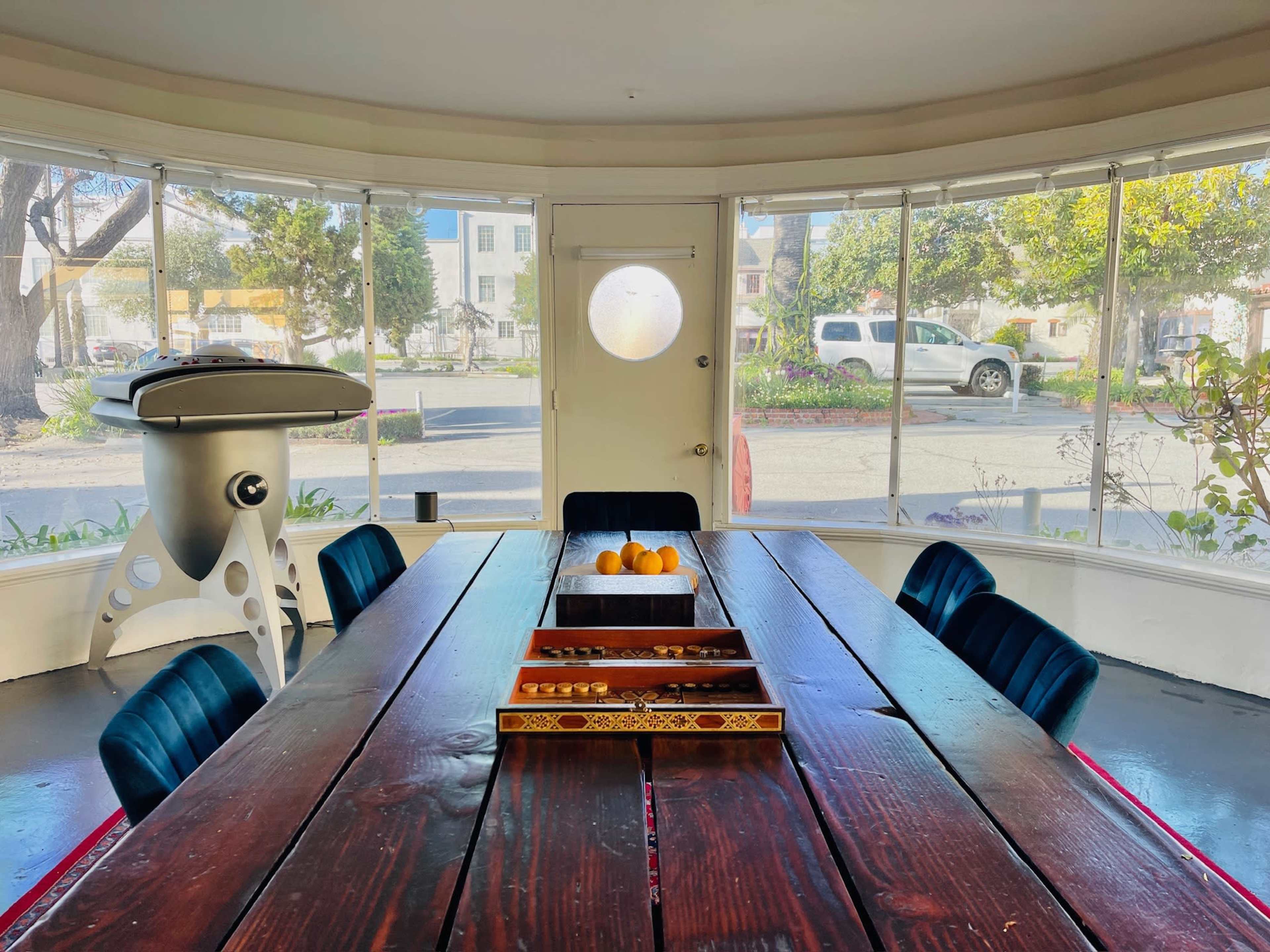 A large wooden dining table with a decorative tray of fruits is set in a bright room surrounded by large windows.