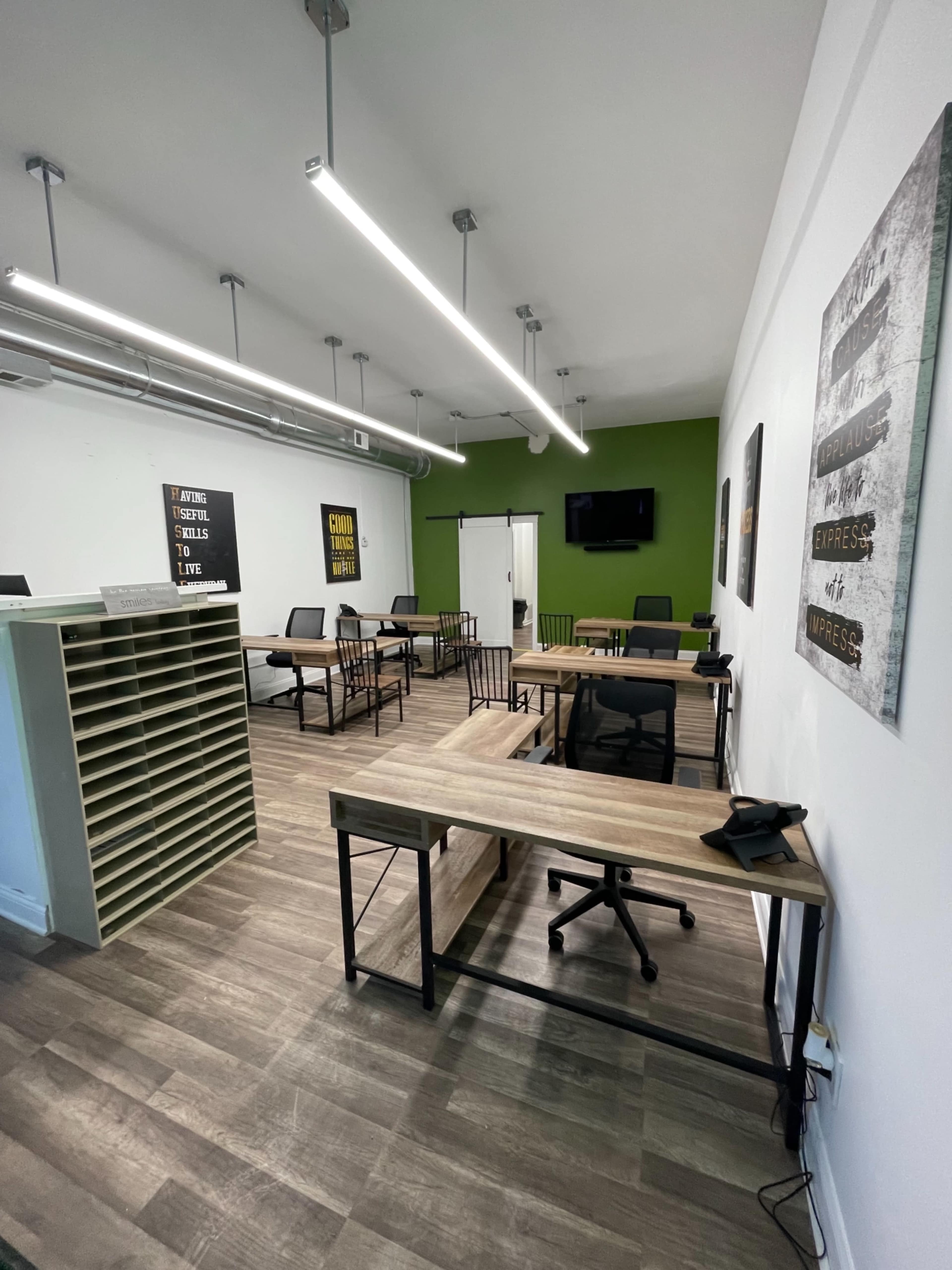 A modern office space with several desks, chairs, and a green accent wall, featuring both shelving and a television mounted on the wall.
