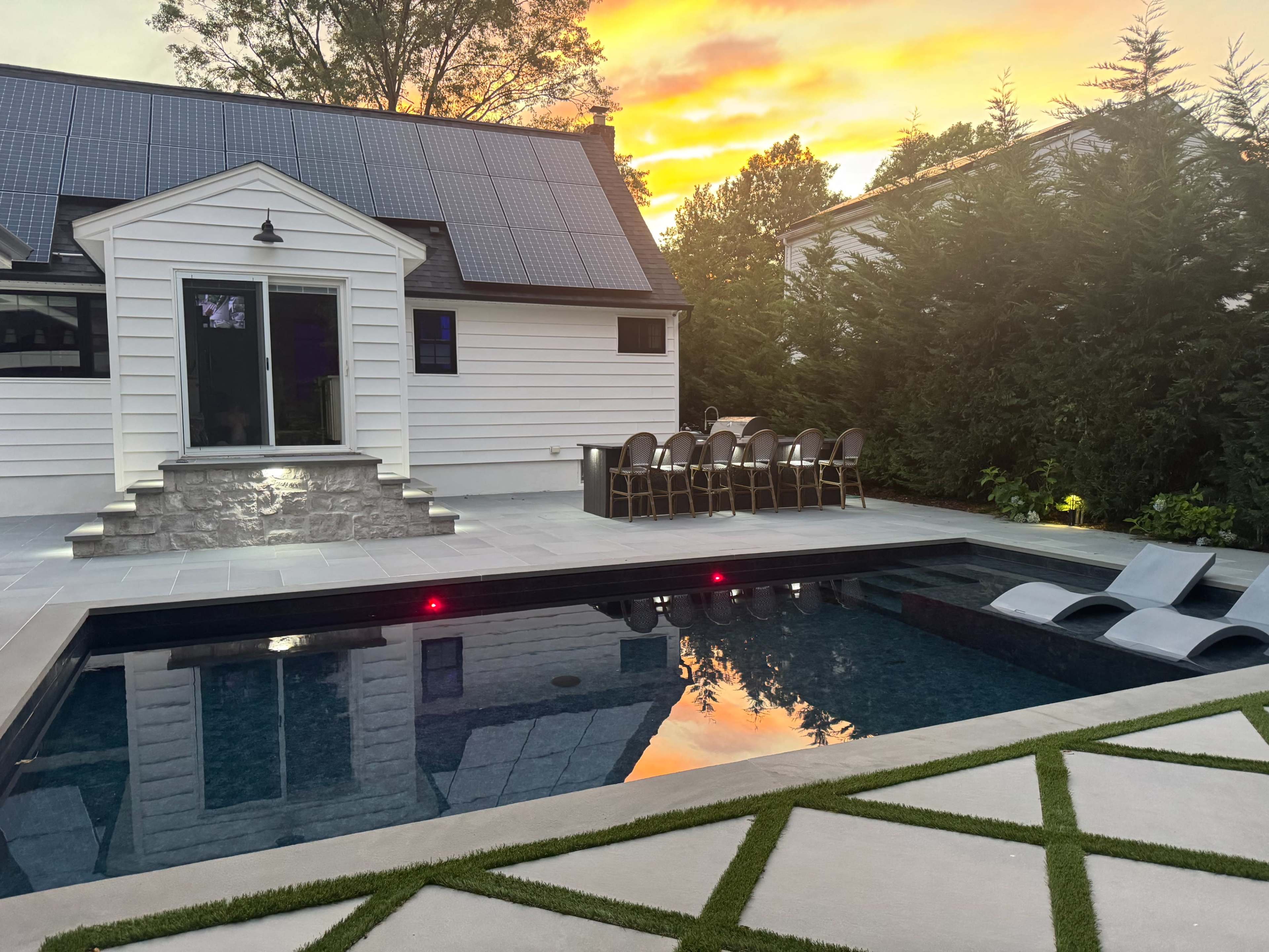 The image shows a modern backyard with a swimming pool, lounge chairs, and an outdoor dining area, all set against a vibrant sunset.