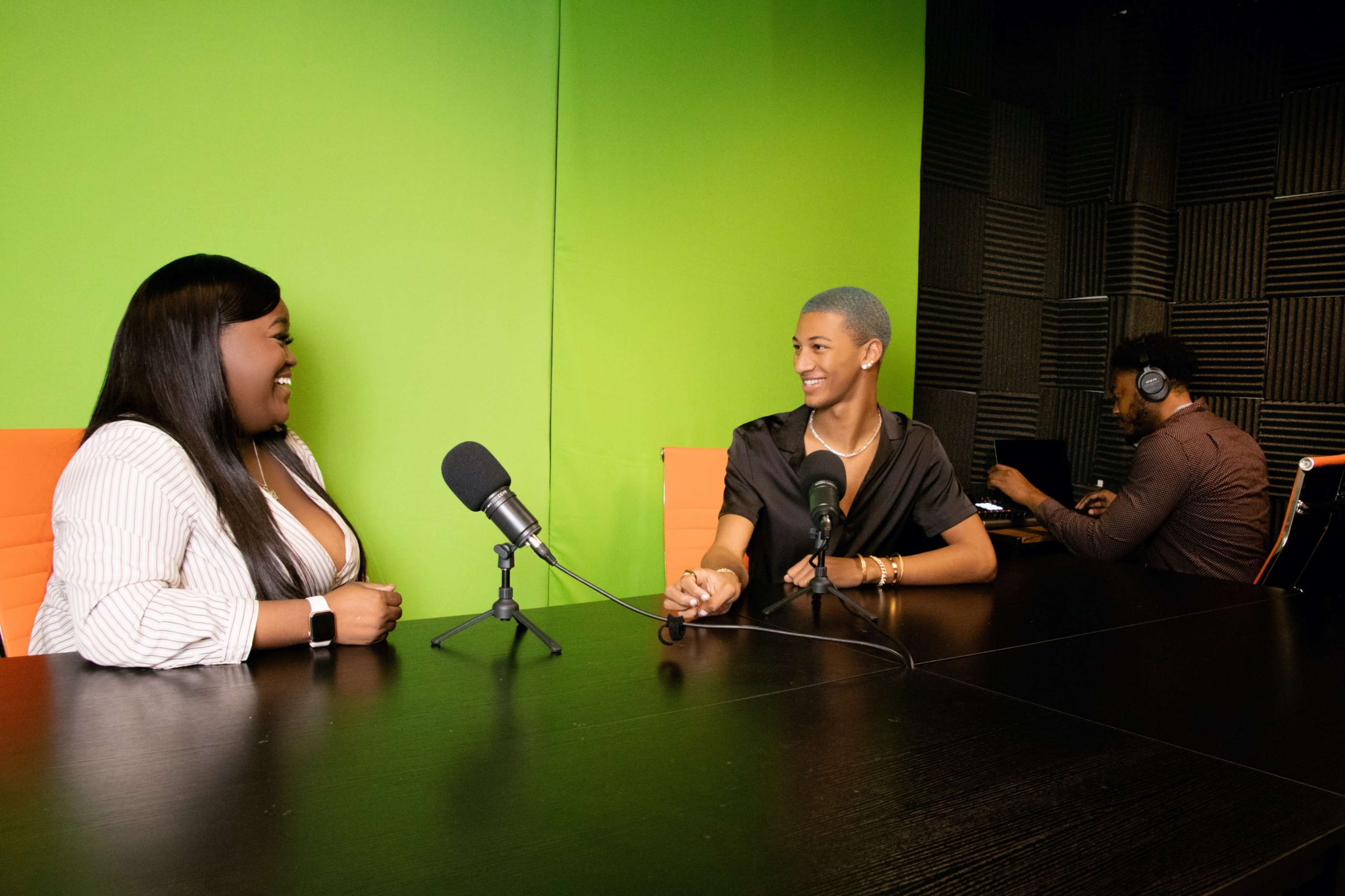 Two women are seated at a table with microphones in front of them, laughing and engaged in conversation, while a man in the background is working at a computer, all set against a green screen backdrop.
