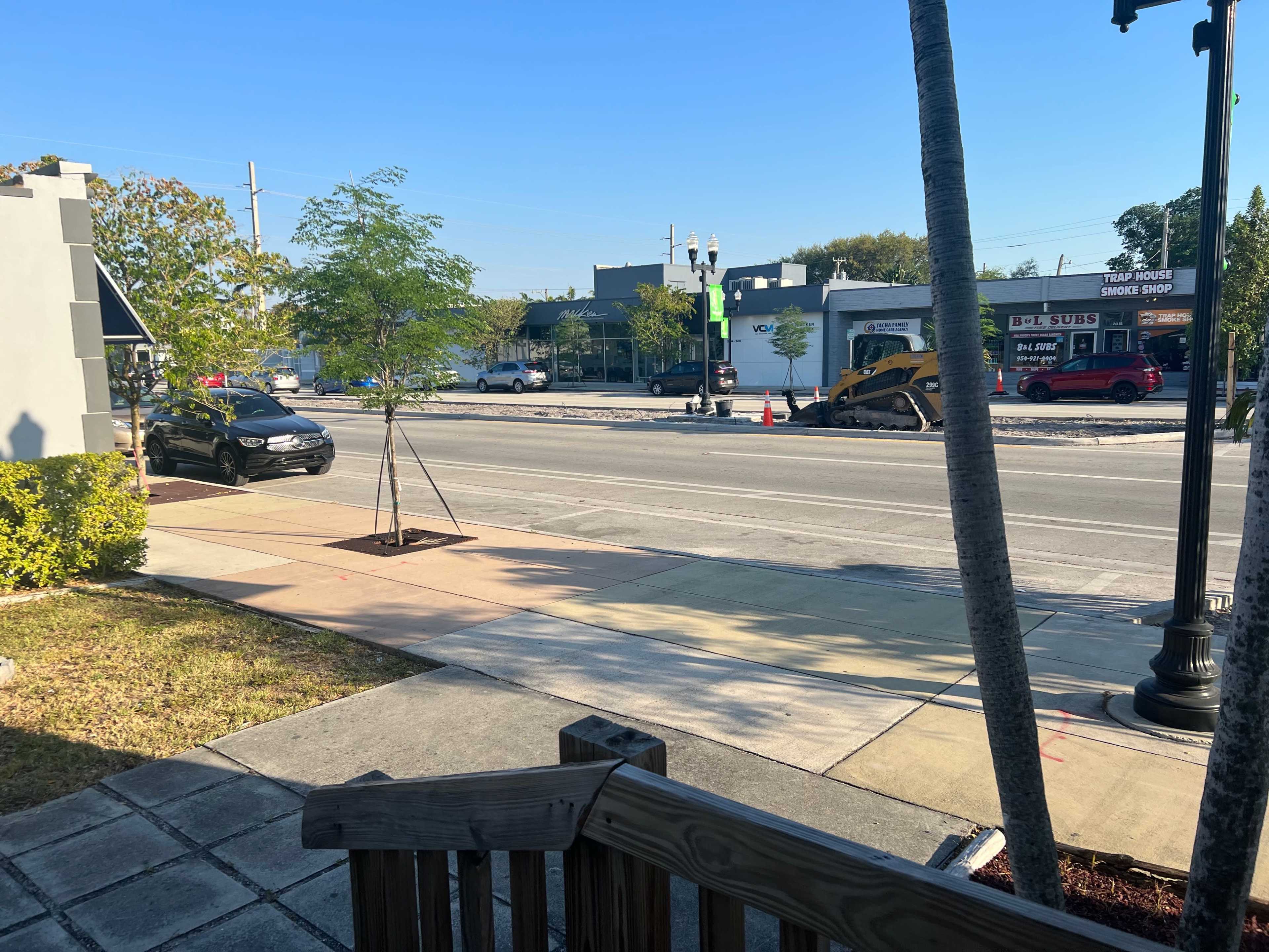 The image shows a street scene with a few parked cars, construction equipment, and commercial buildings in the background under a clear blue sky.