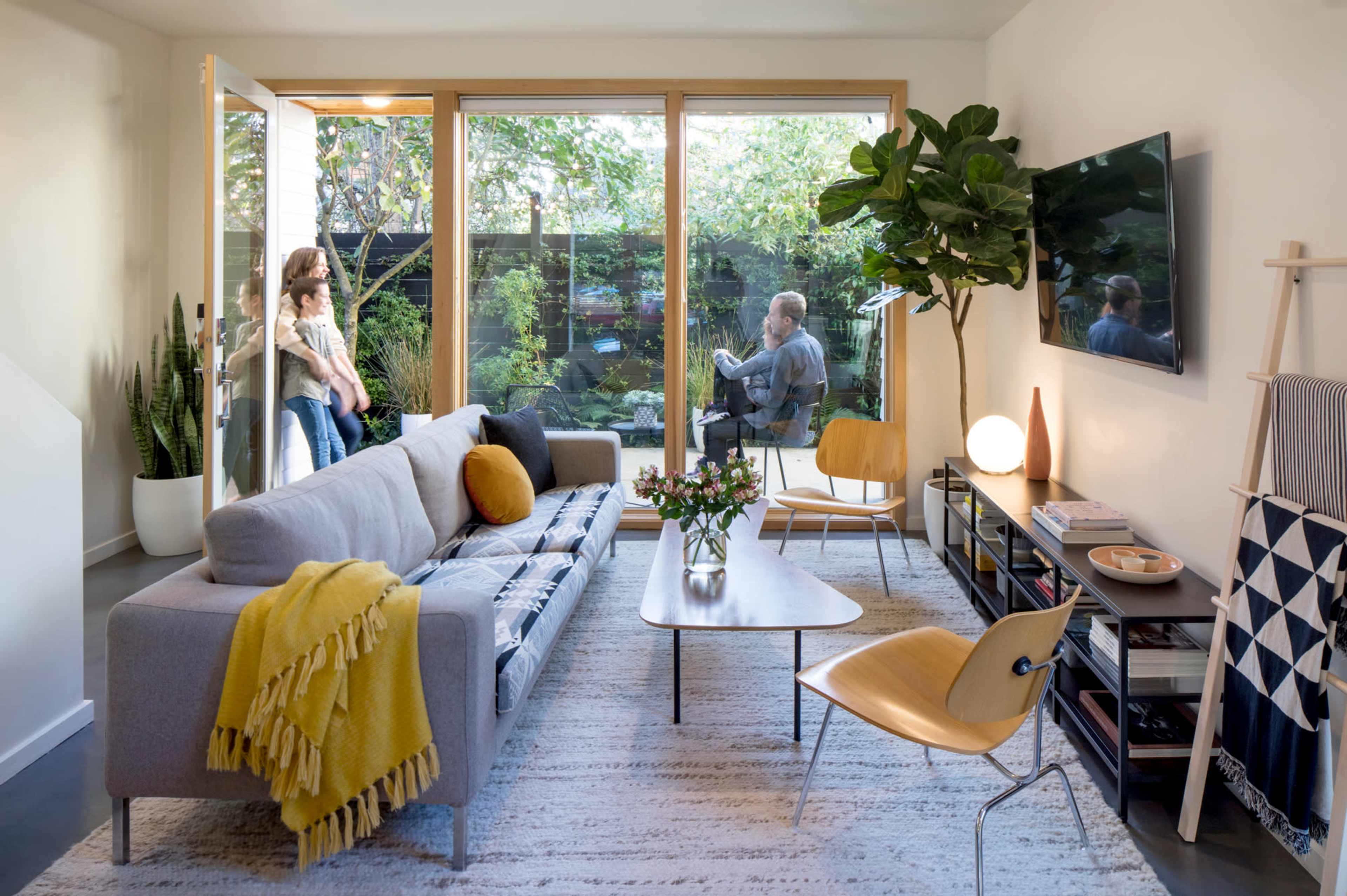A modern living room features a gray sofa with a yellow throw, a coffee table, and a large window opening to a garden, while a person sits outside and two others stand inside.