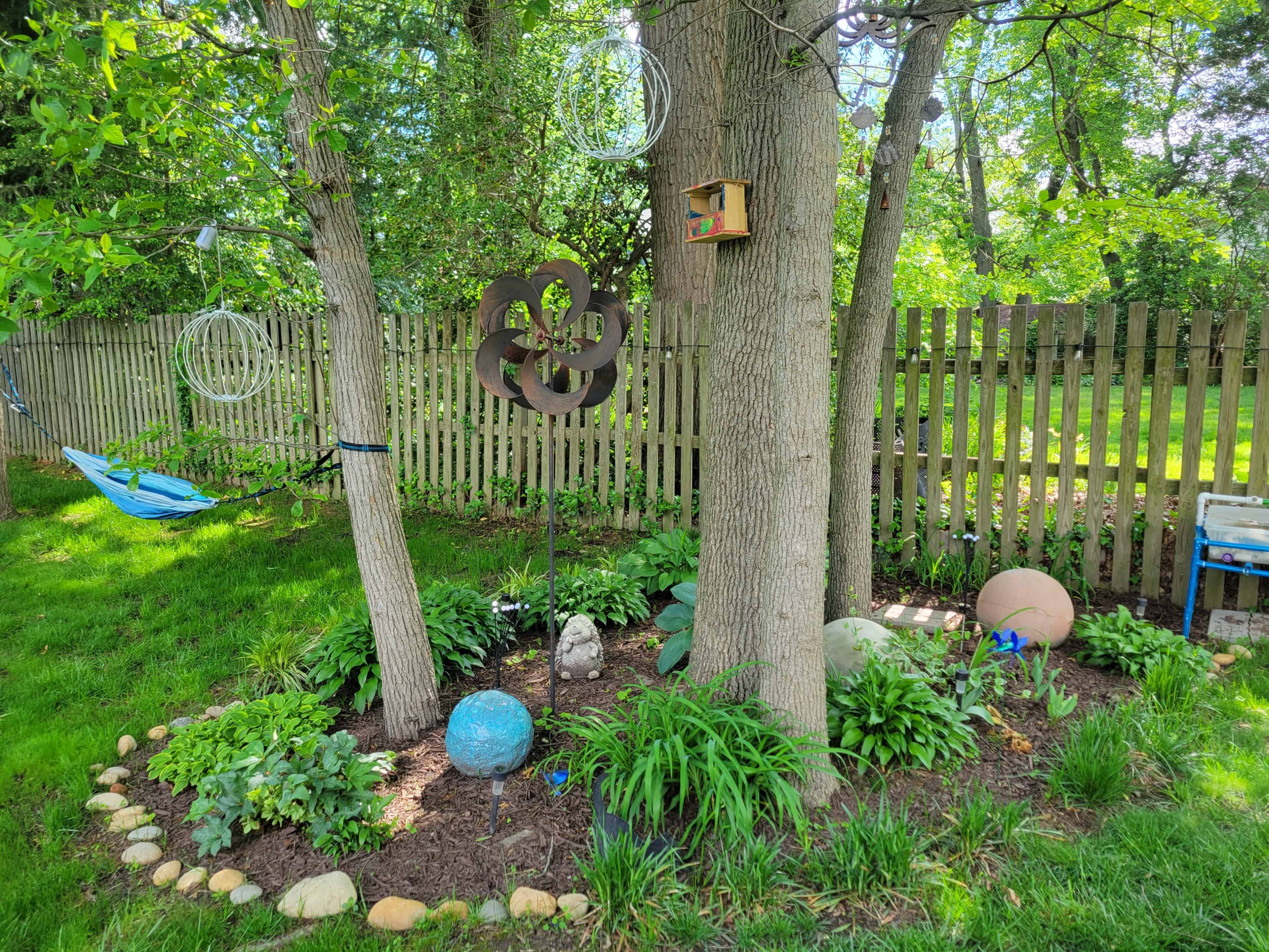 The image depicts a landscaped garden area between two trees, featuring a metal sculpture, colorful stones, plants, and a birdhouse near a wooden fence.