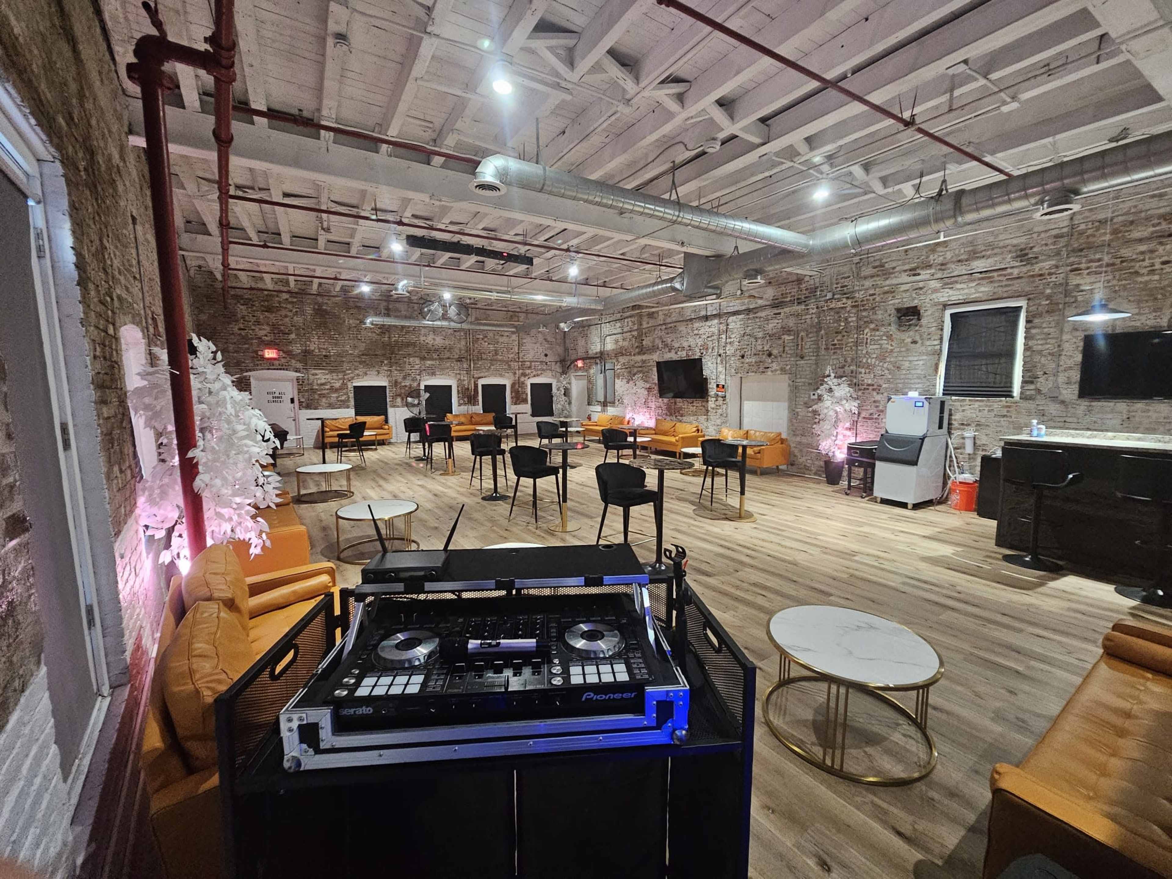 A spacious event venue with brick walls, wooden floors, and seating areas equipped with tables and chairs, illuminated by soft lighting.