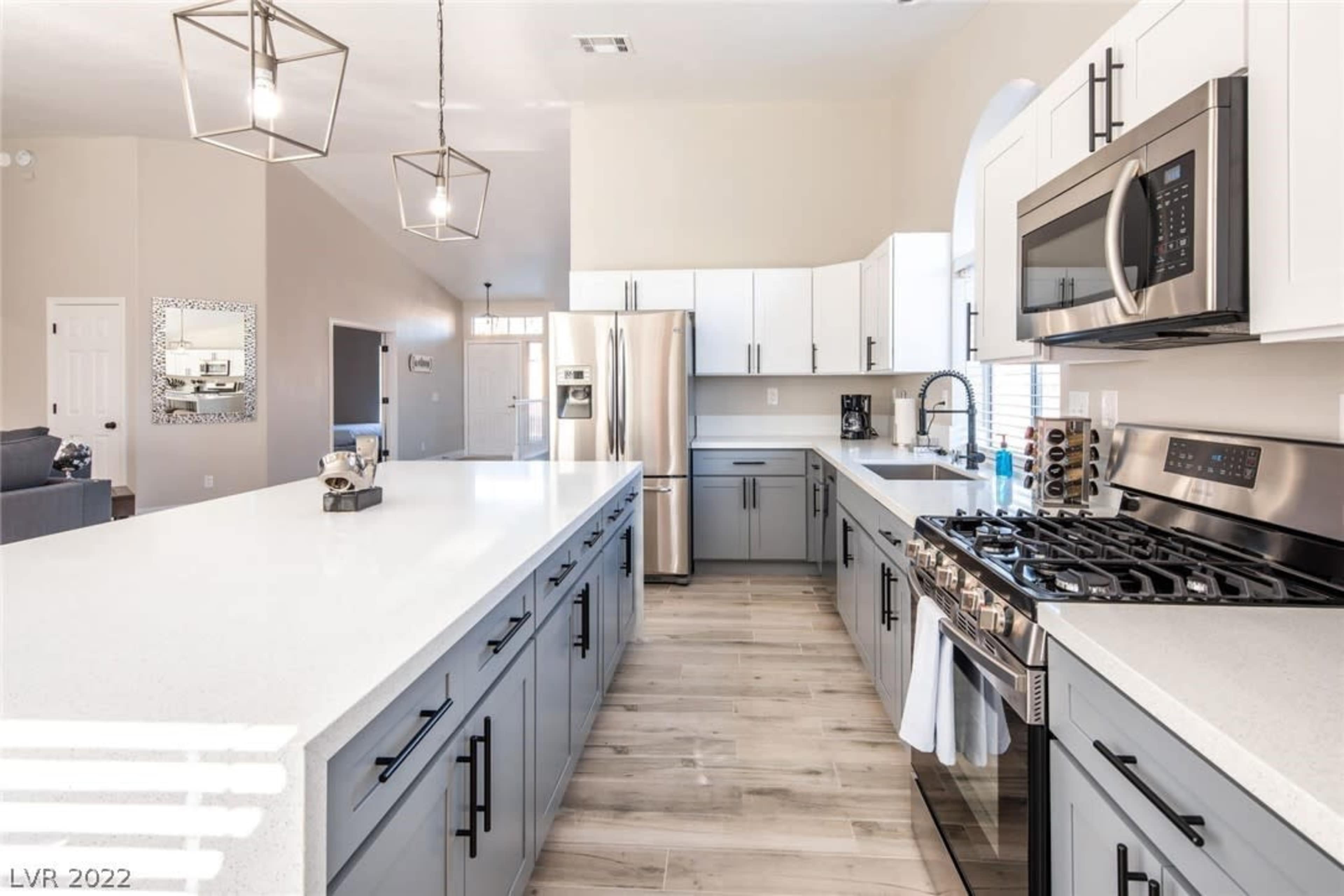 The image shows a modern kitchen with white and gray cabinetry, stainless steel appliances, and a large island with a white countertop.