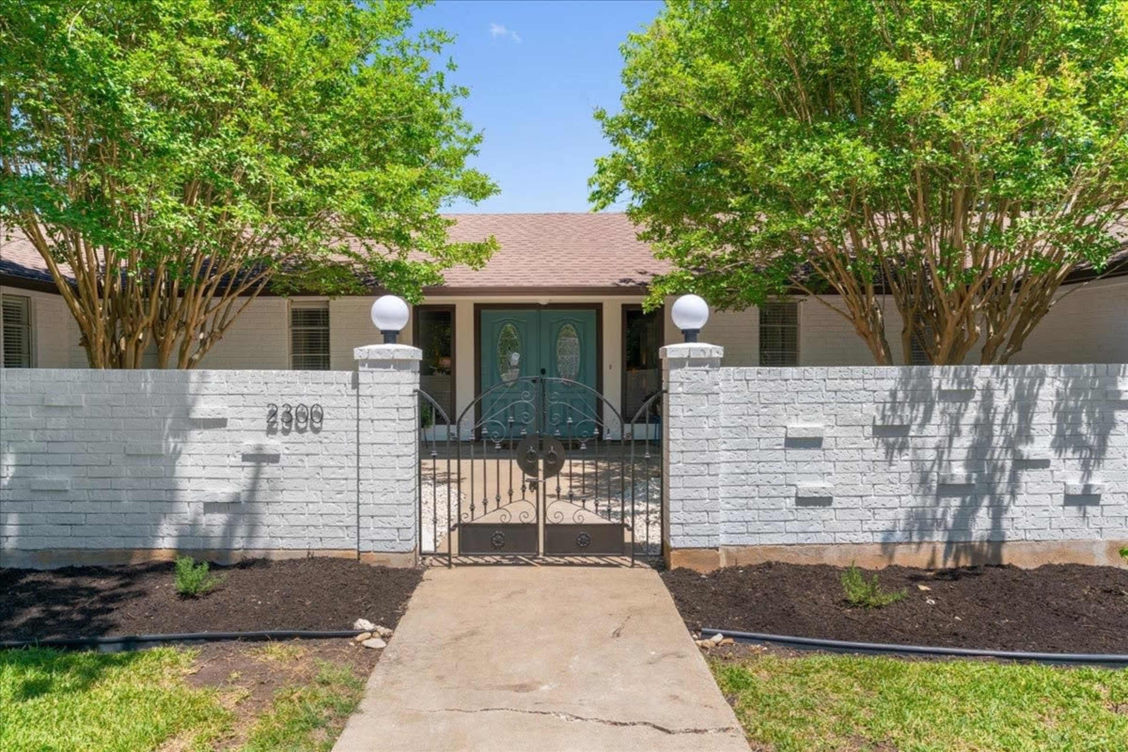 A front yard with a white brick wall and gate leading to a house with green doors, surrounded by trees.