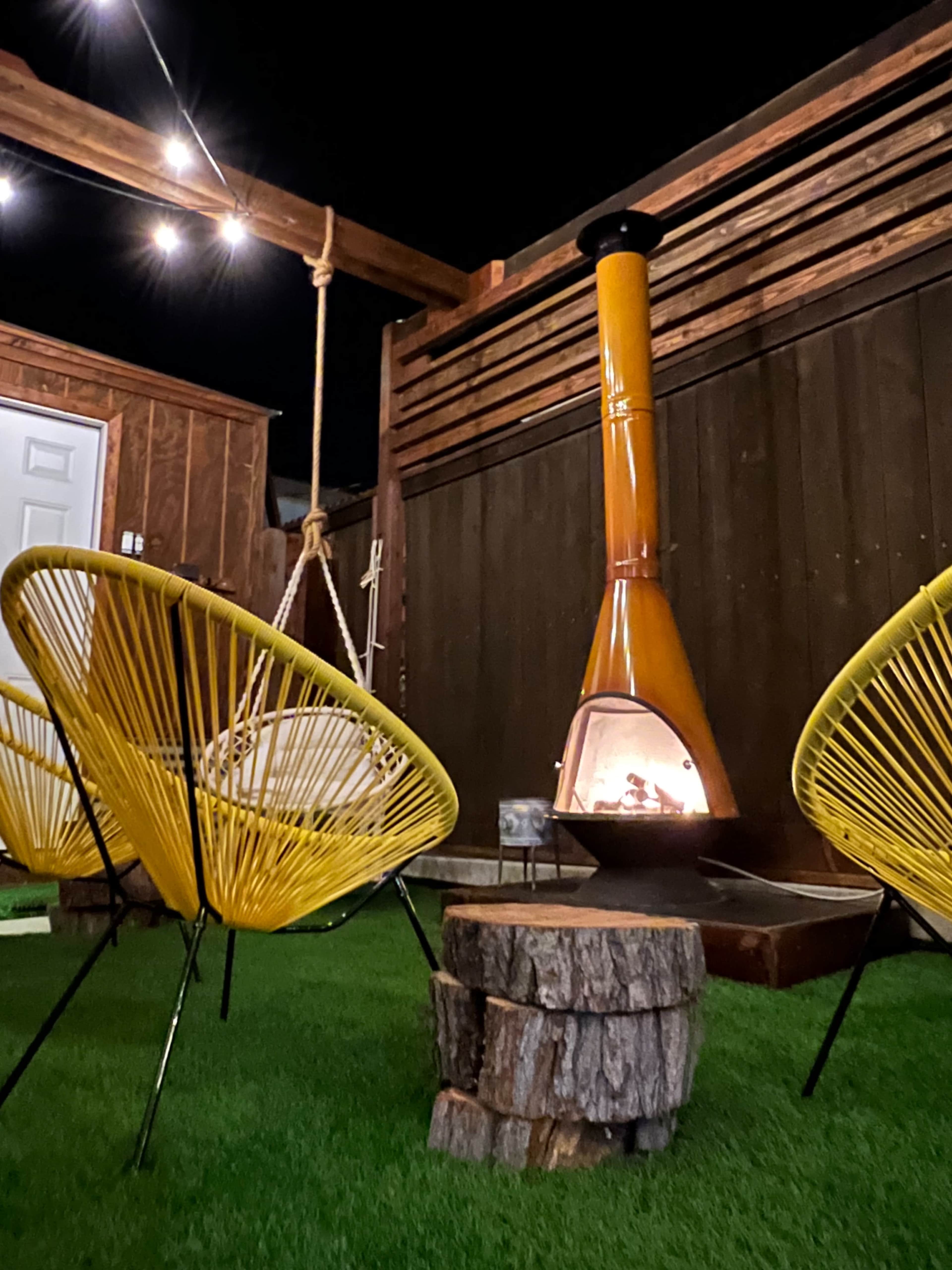 A cozy outdoor space features two yellow chairs facing a chiminea on a wooden platform with green artificial grass.