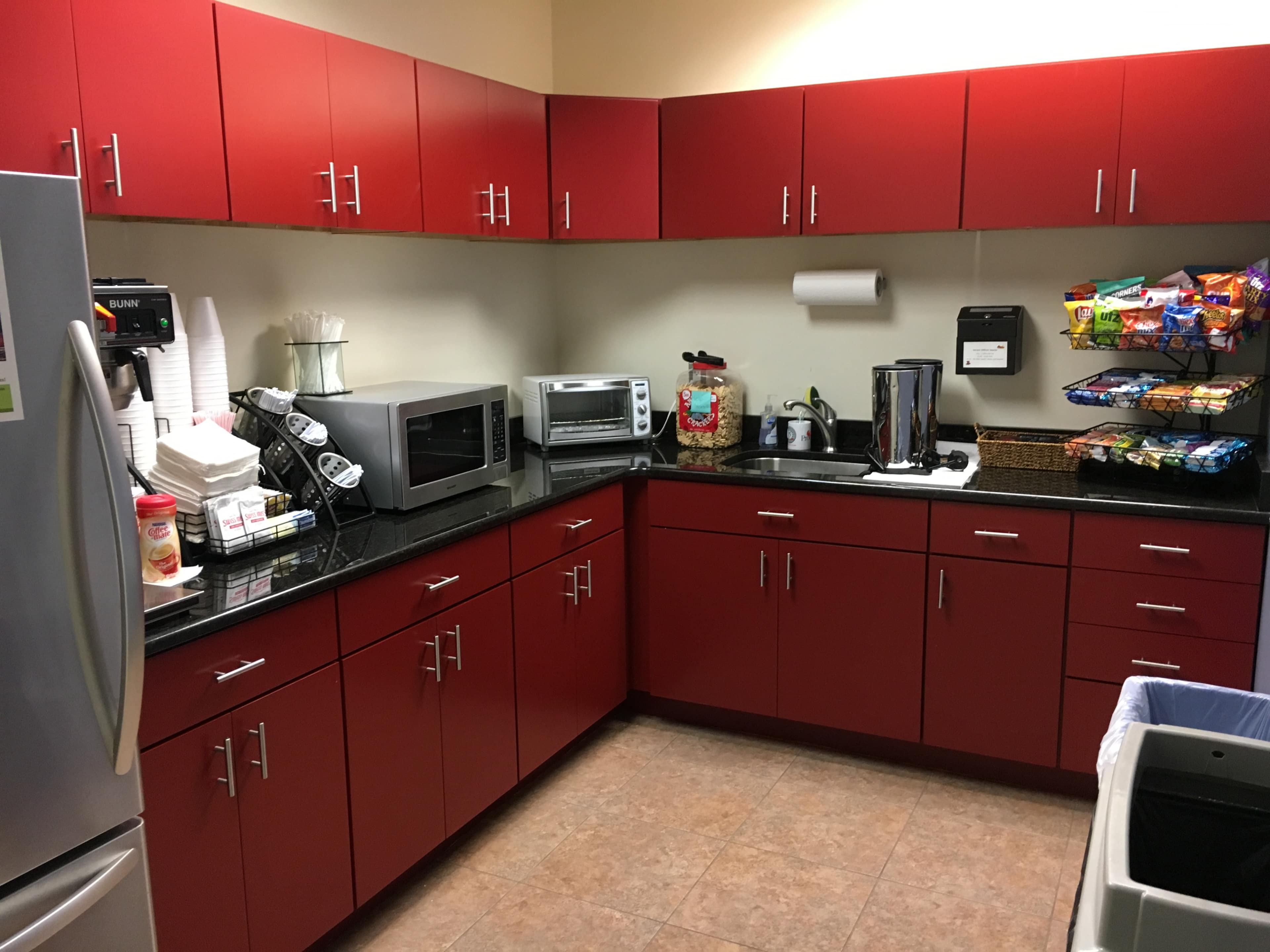 A kitchen with red cabinets, a countertop with a microwave, a coffee maker, and a snack rack on the wall.