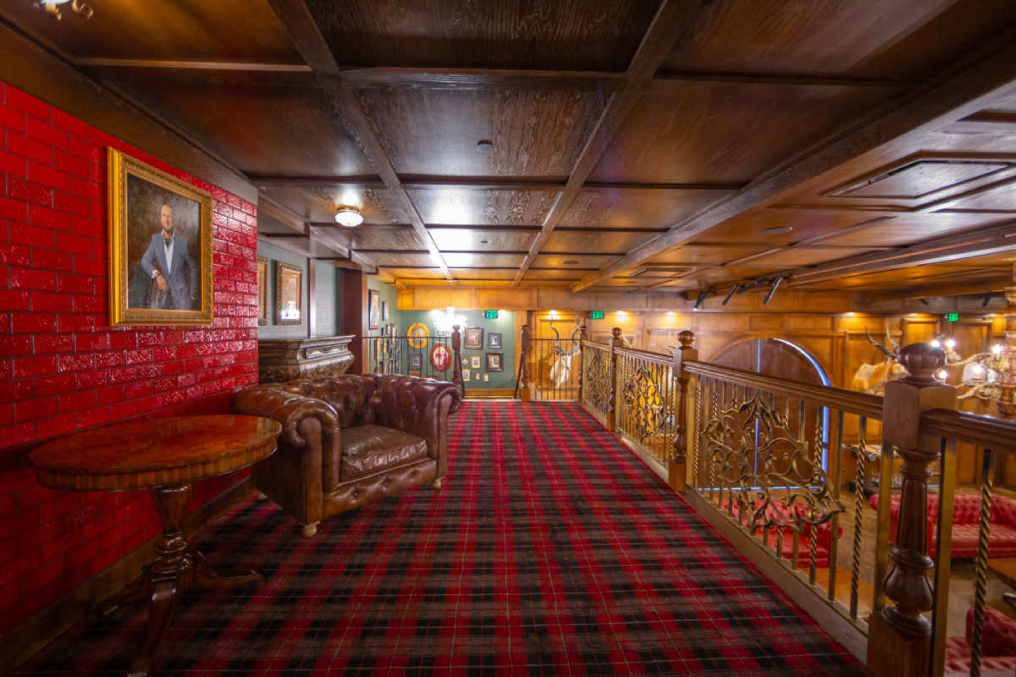 The image shows a wooden interior hallway featuring a red-brick wall, a leather couch, a round table, and framed artwork.