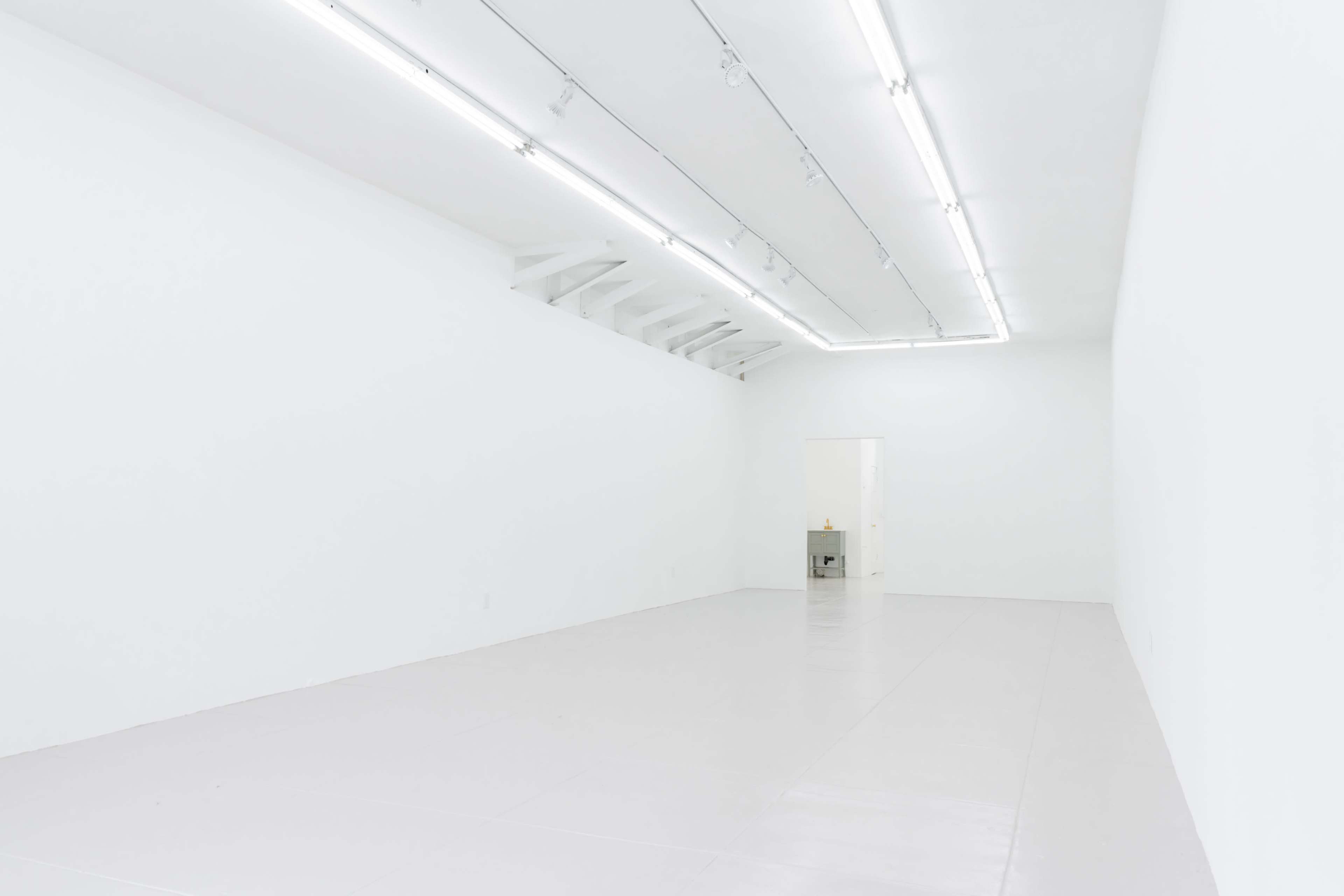 The image depicts a minimalistic, empty white gallery space with bright overhead lighting and a door in the back.