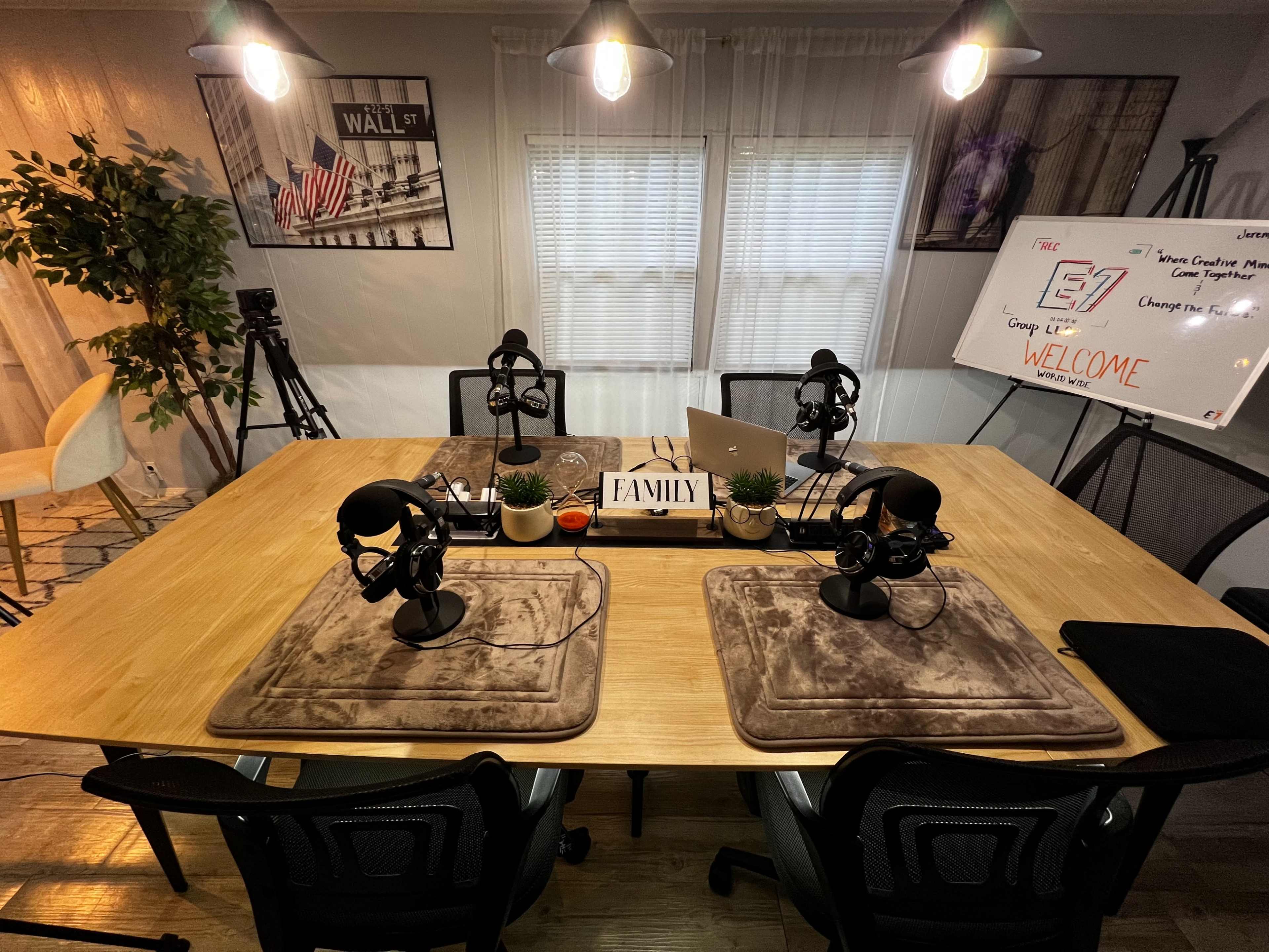 A recording studio with a large wooden table, multiple microphones, laptops, and a whiteboard set up for a podcast or discussion, surrounded by plants and studio lights.