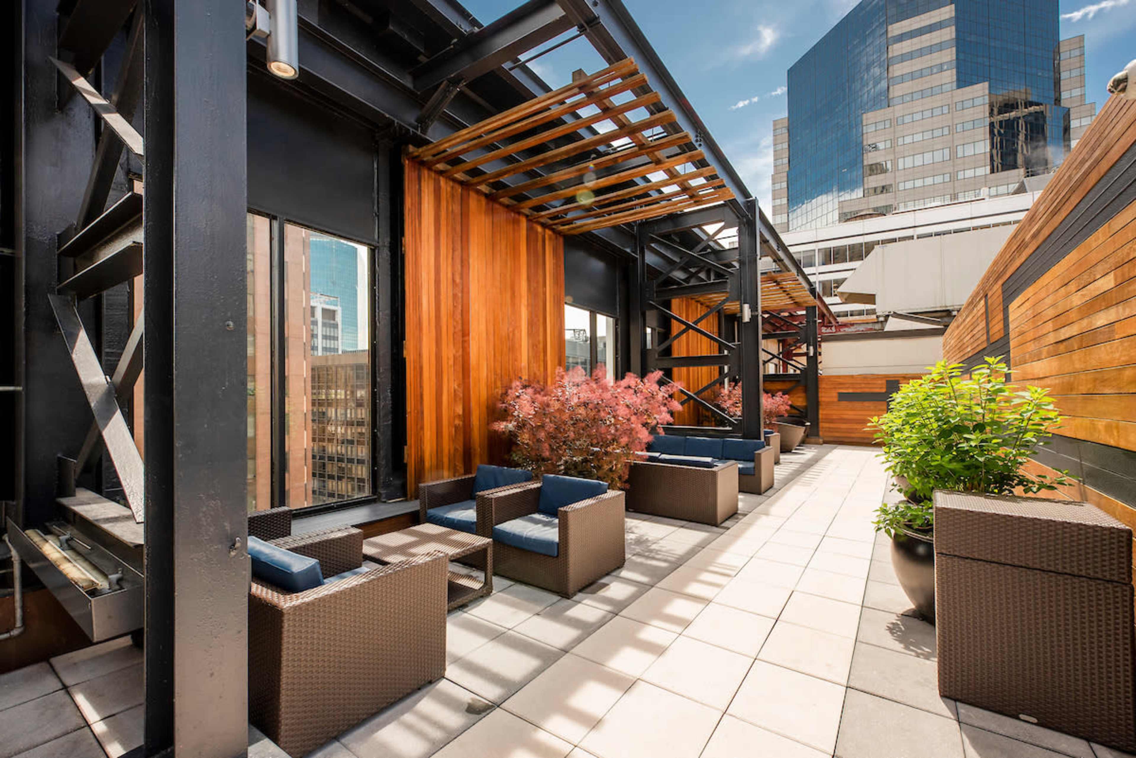 A modern rooftop terrace features woven furniture, wooden panels, and city skyline views.