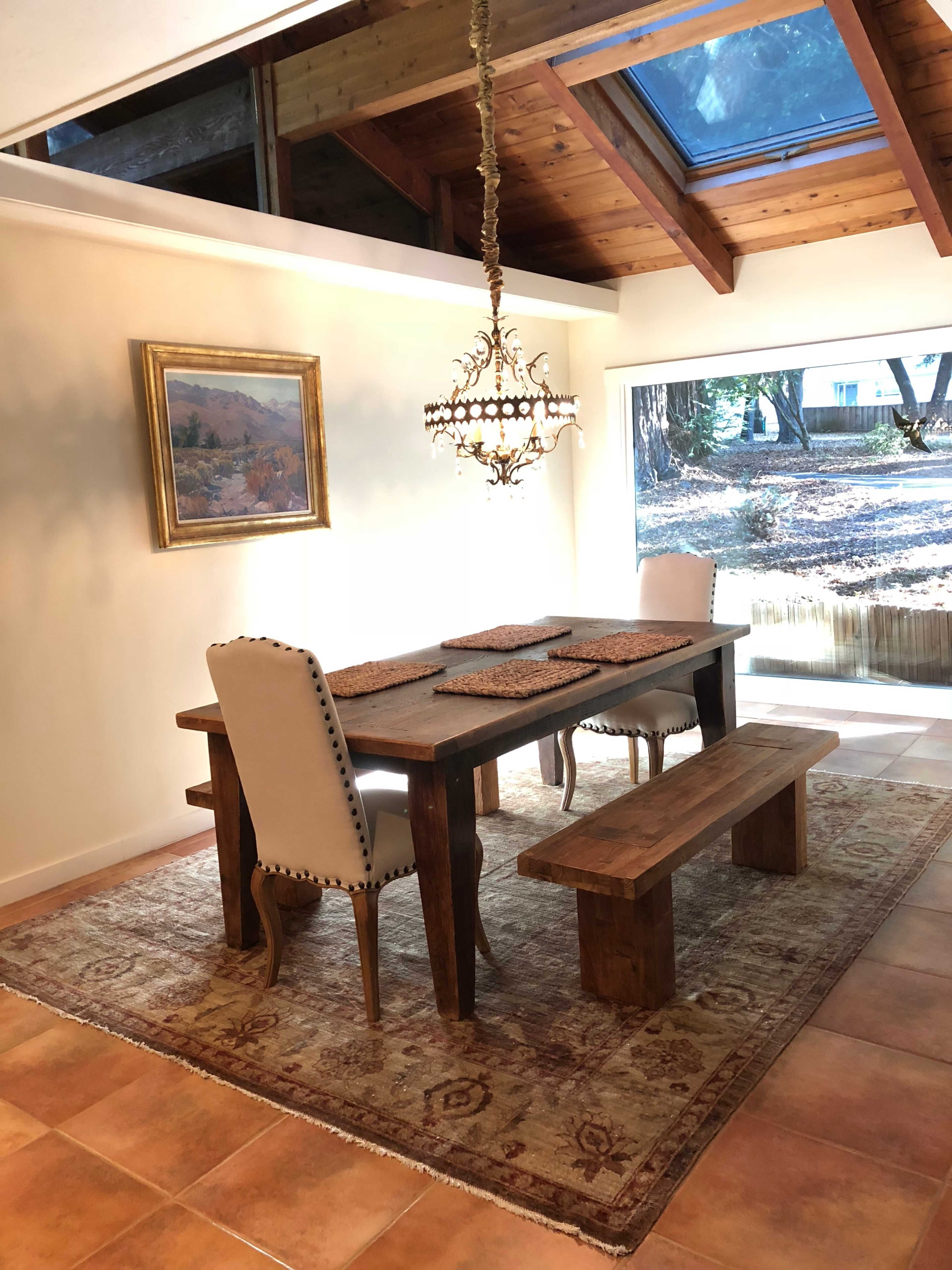 A dining area with a wooden table, two upholstered chairs, a bench, decorative placemats, and a chandelier under a slanted ceiling with a skylight.