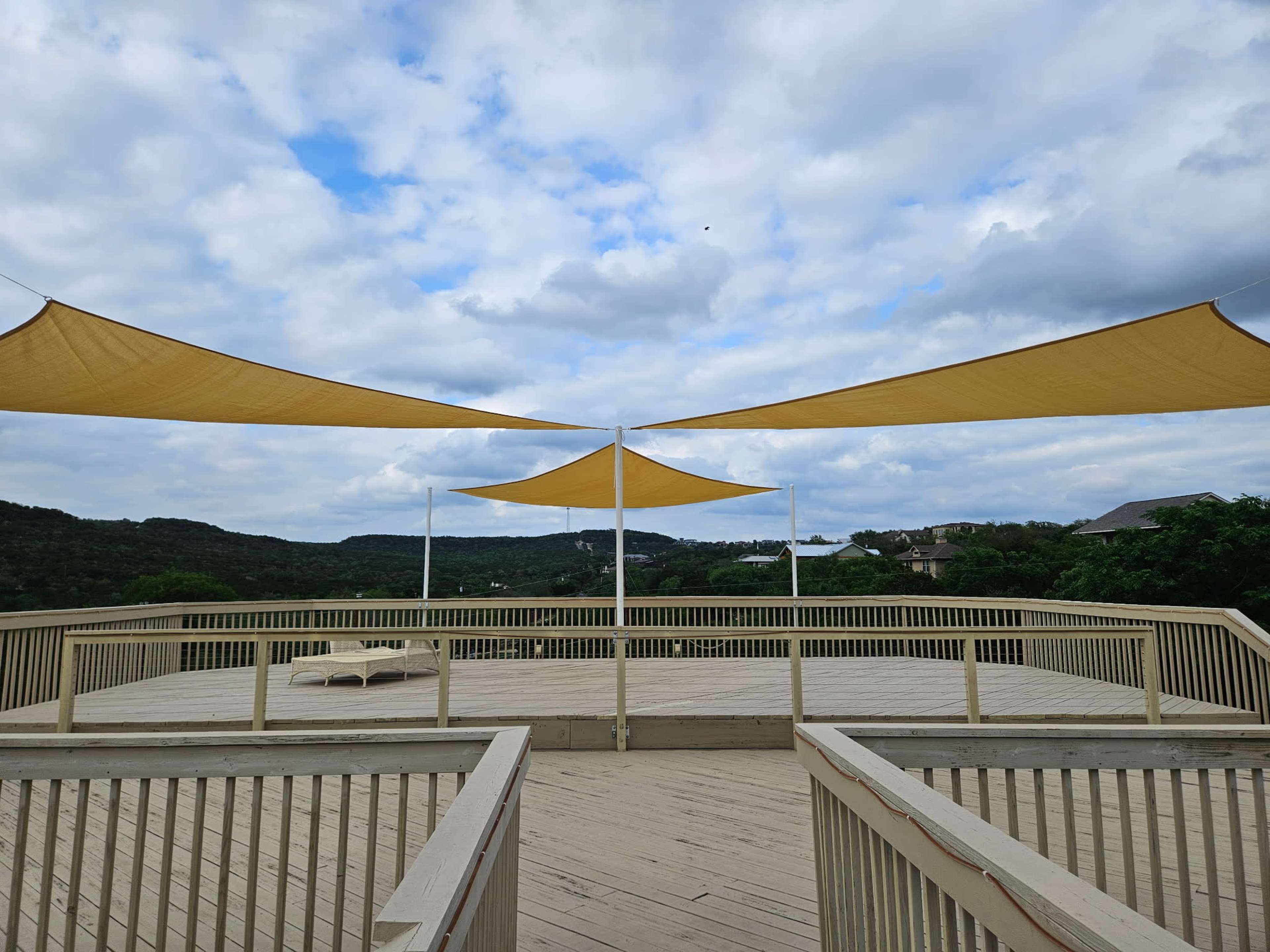 A wooden deck featuring two yellow shade sails stretches towards a hilly landscape under a partly cloudy sky.