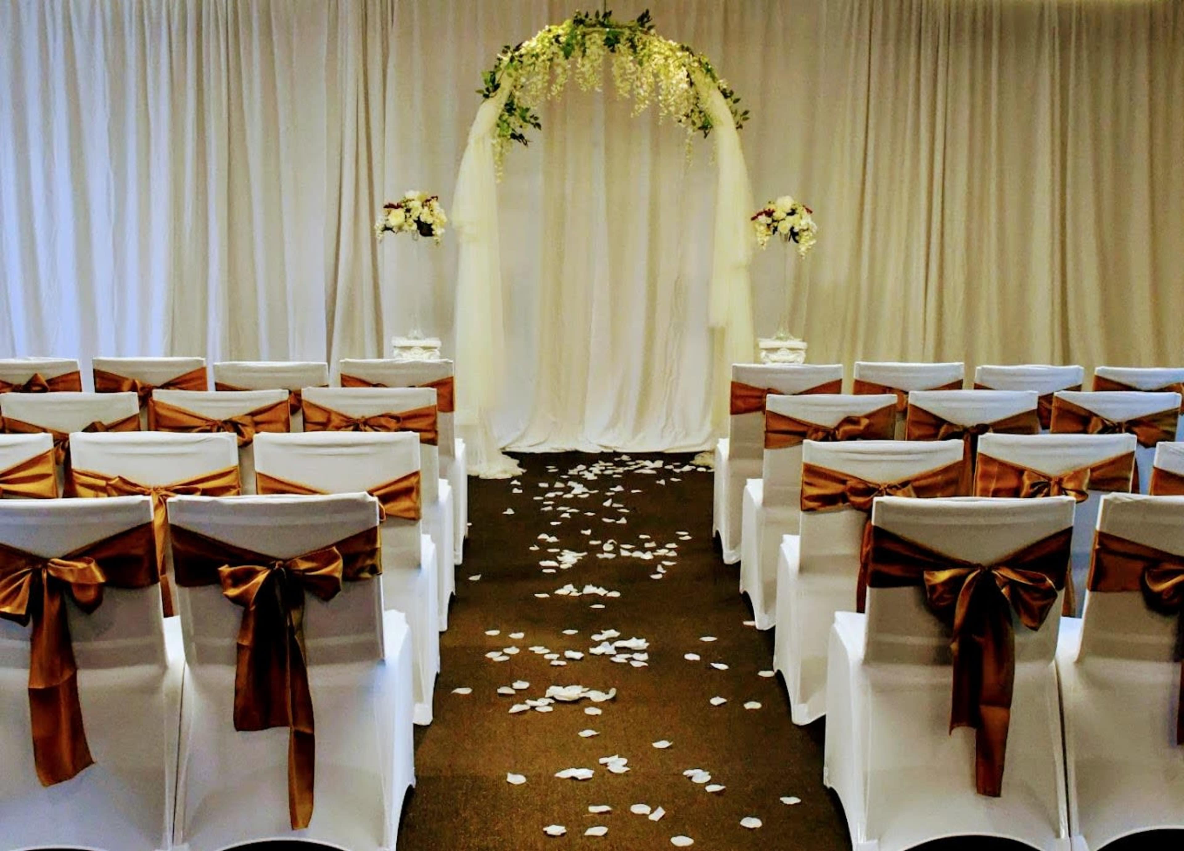 A wedding ceremony setup with white chairs adorned with brown sashes, facing an arch decorated with greenery and flowers, set against a draped backdrop.