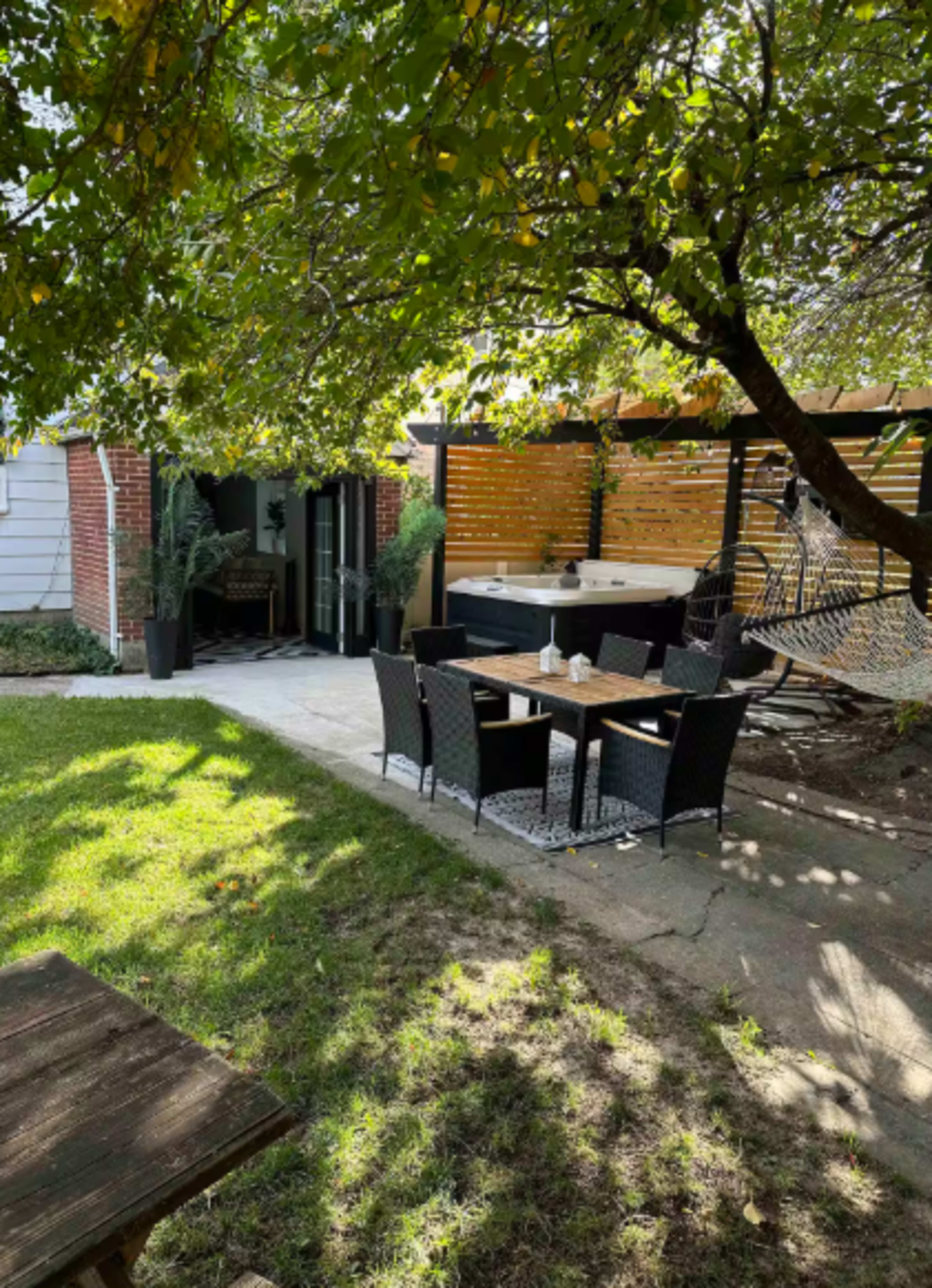 The image shows a backyard area with a seating arrangement, a hot tub, and a hammock beneath trees, surrounded by greenery and a brick exterior on one side.