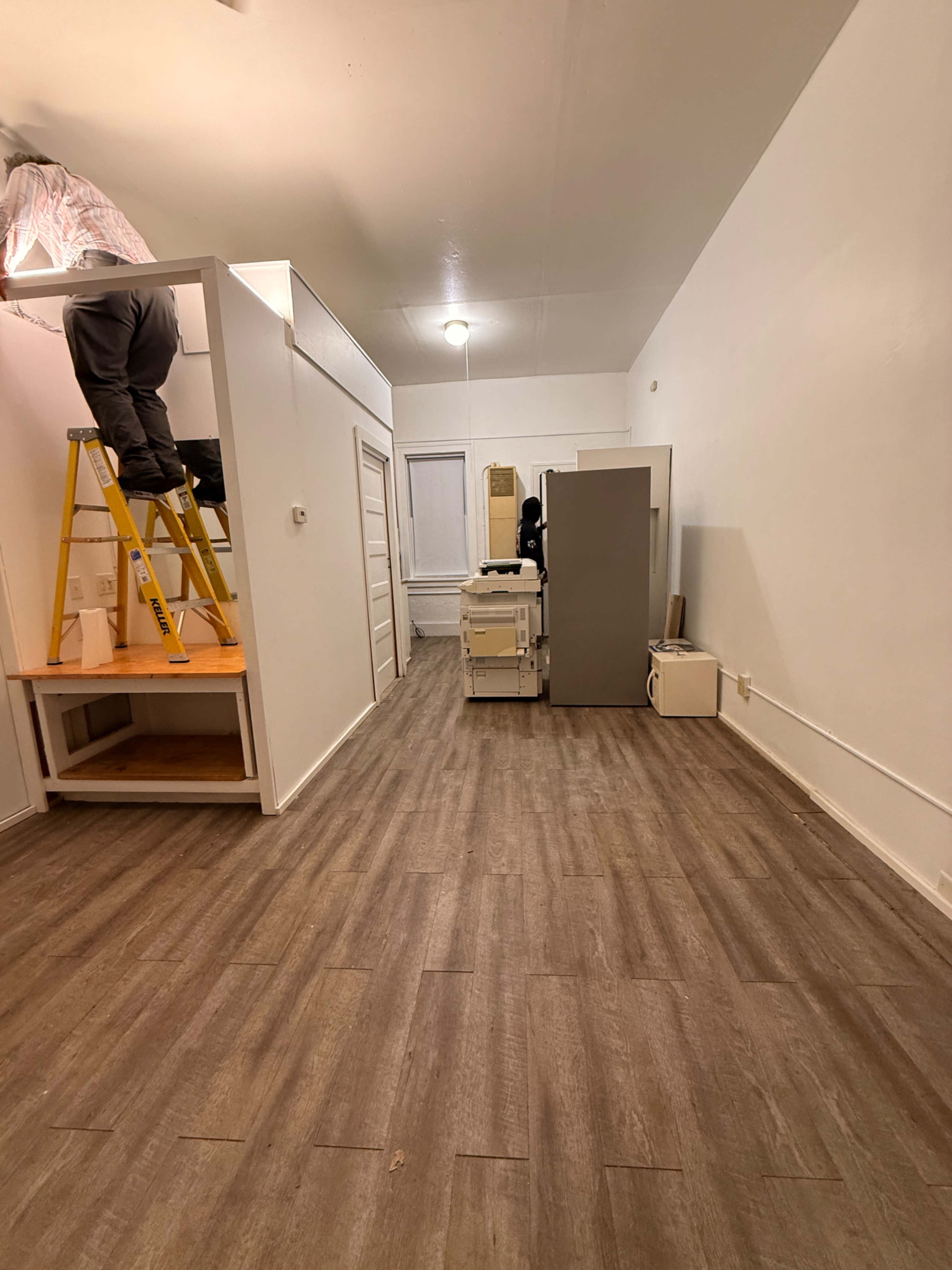 The image shows a spacious room with light-colored walls, a ladder against the wall, a kitchen area at the left, and a gray refrigerator and printer in the center.