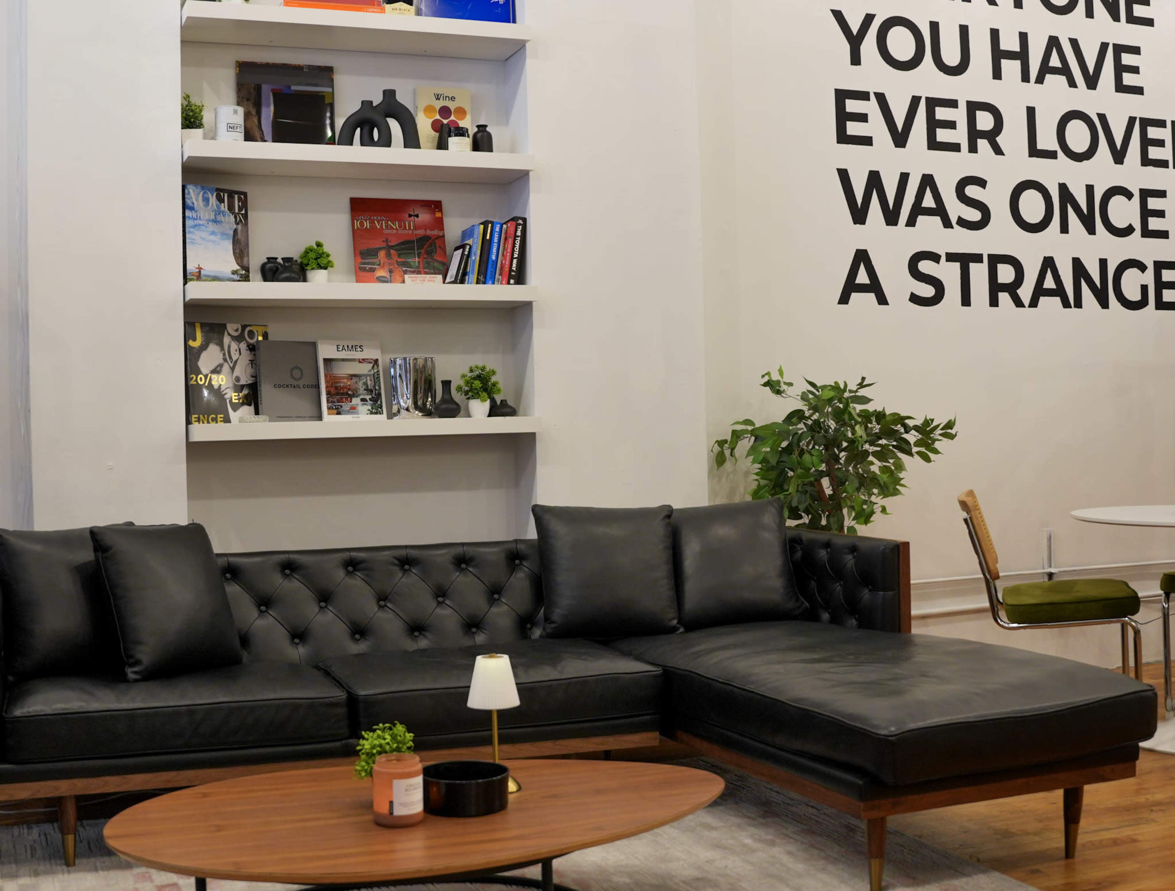 A modern lounge area features a black leather sectional sofa, a wooden coffee table, and a wall with a quote, alongside shelves displaying books and decorative items.