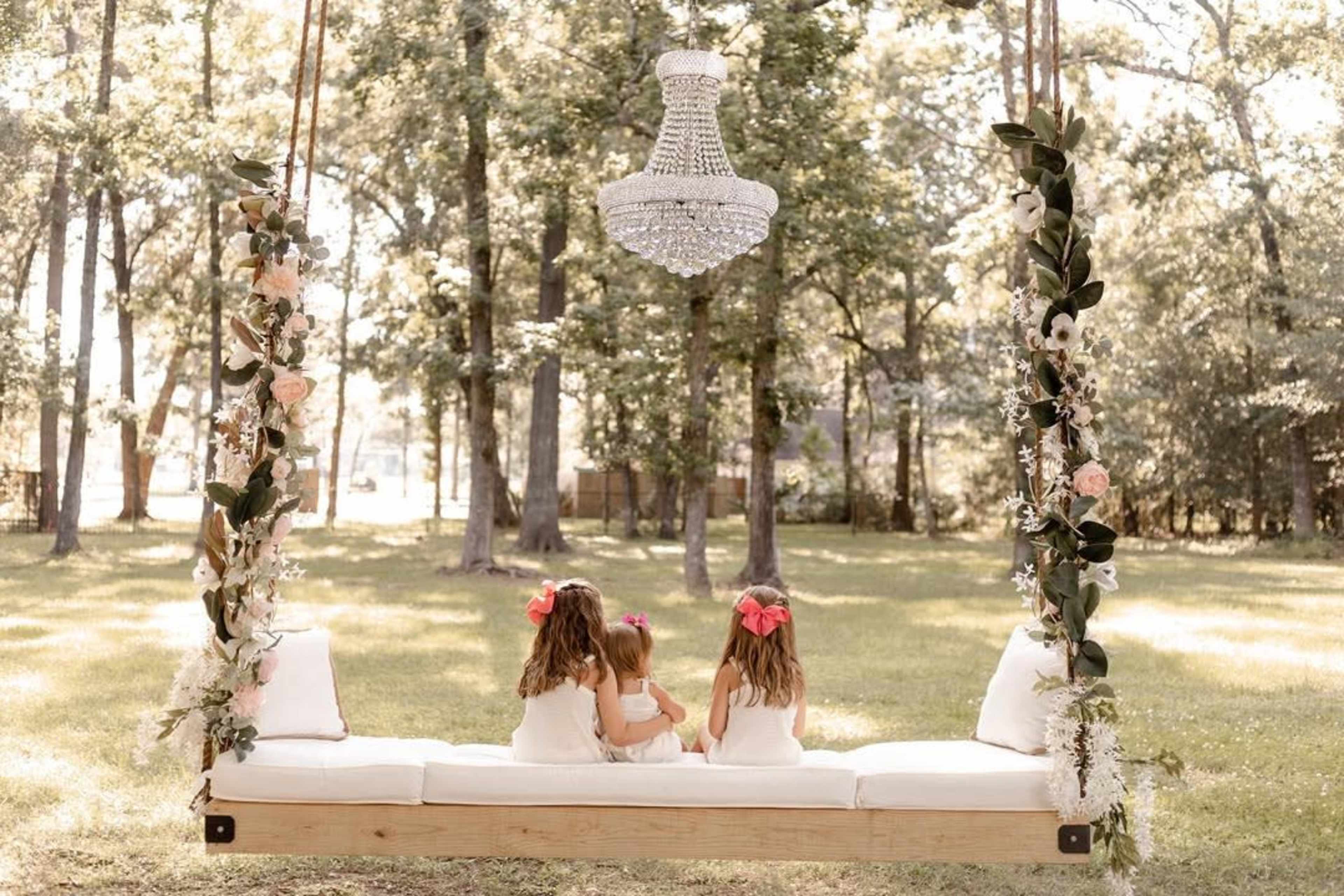 Two children sit on a wooden swing decorated with flowers under a chandelier in a green park.