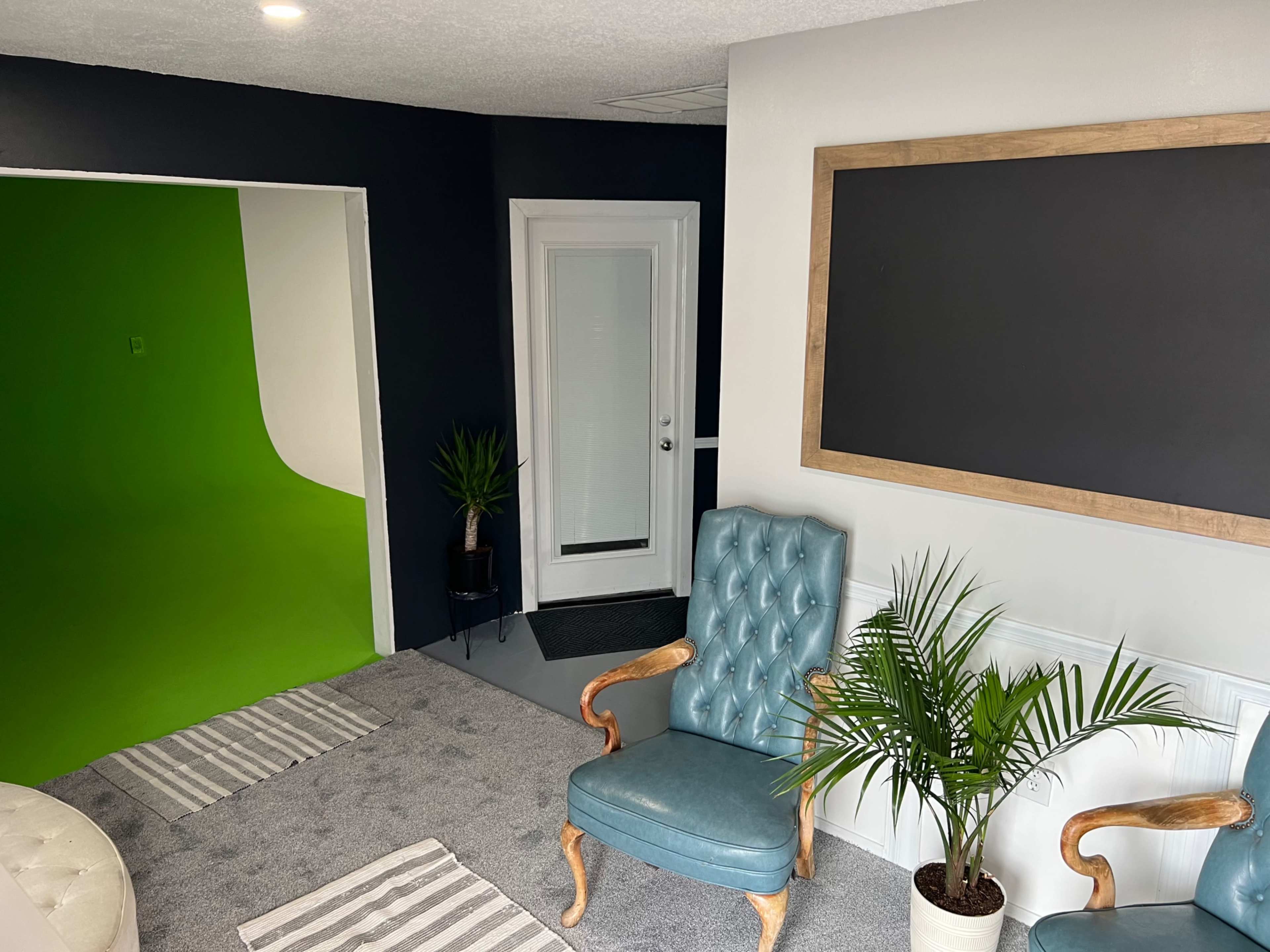 A modern interior space featuring a green screen area, two upholstered chairs, a small plant, and a large wall-mounted chalkboard frame.