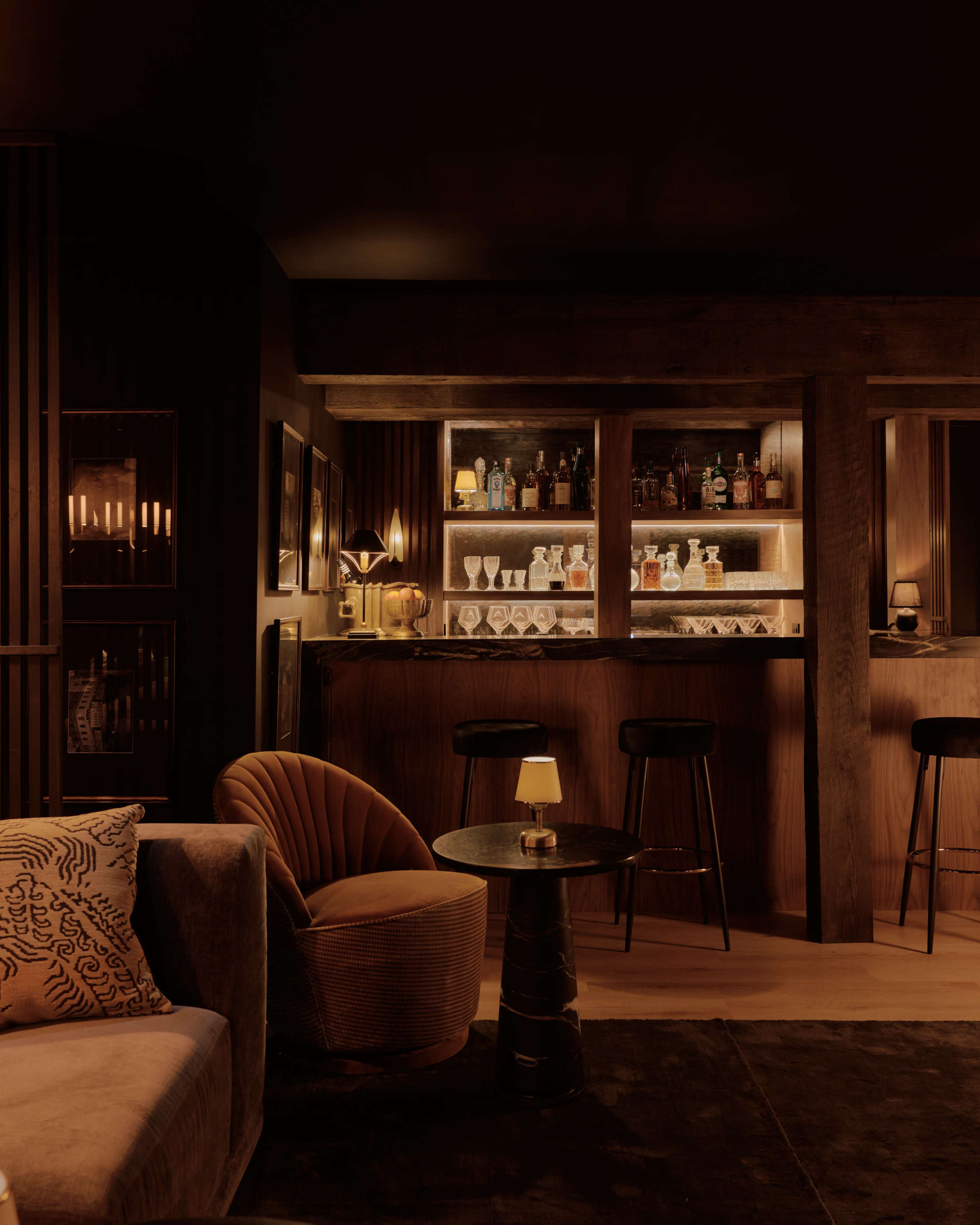 The image shows a dimly lit bar area with a wooden counter, various bottles displayed on shelves, and cozy seating arrangements.