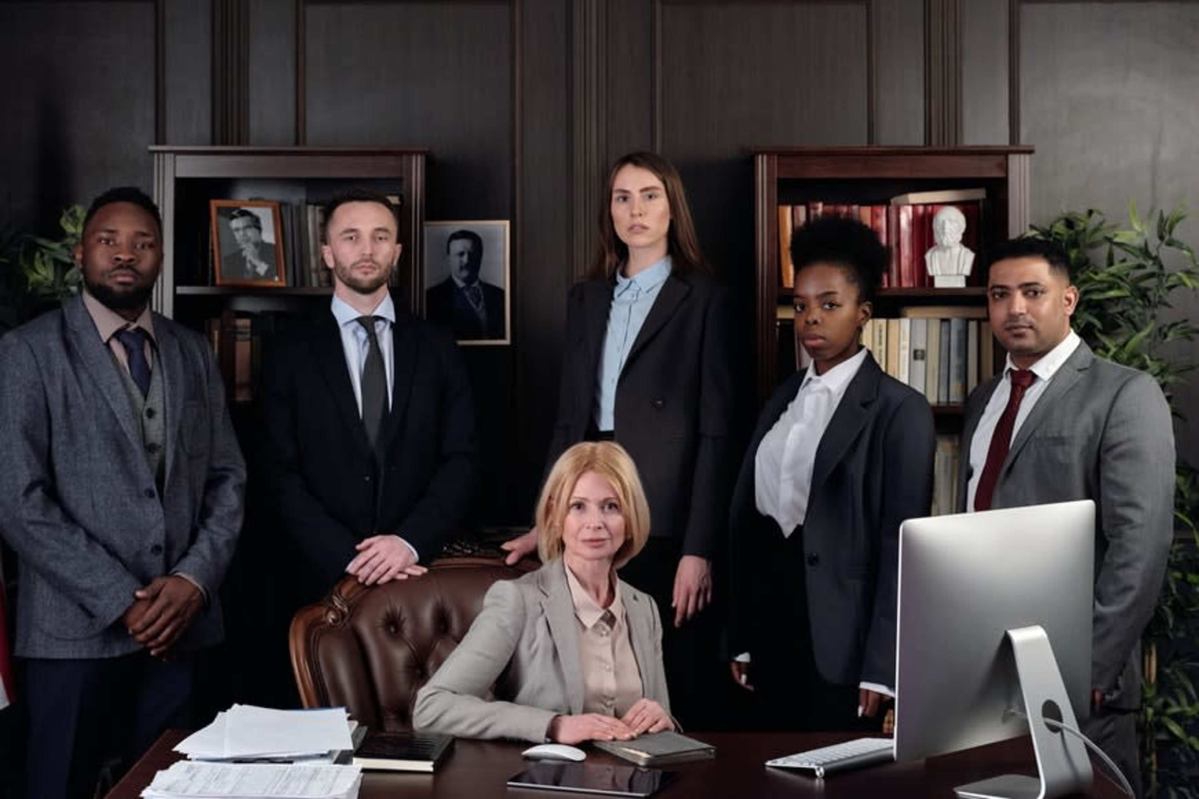 A group of six professionals in business attire stands and sits in a well-furnished office with bookshelves and portraits in the background.