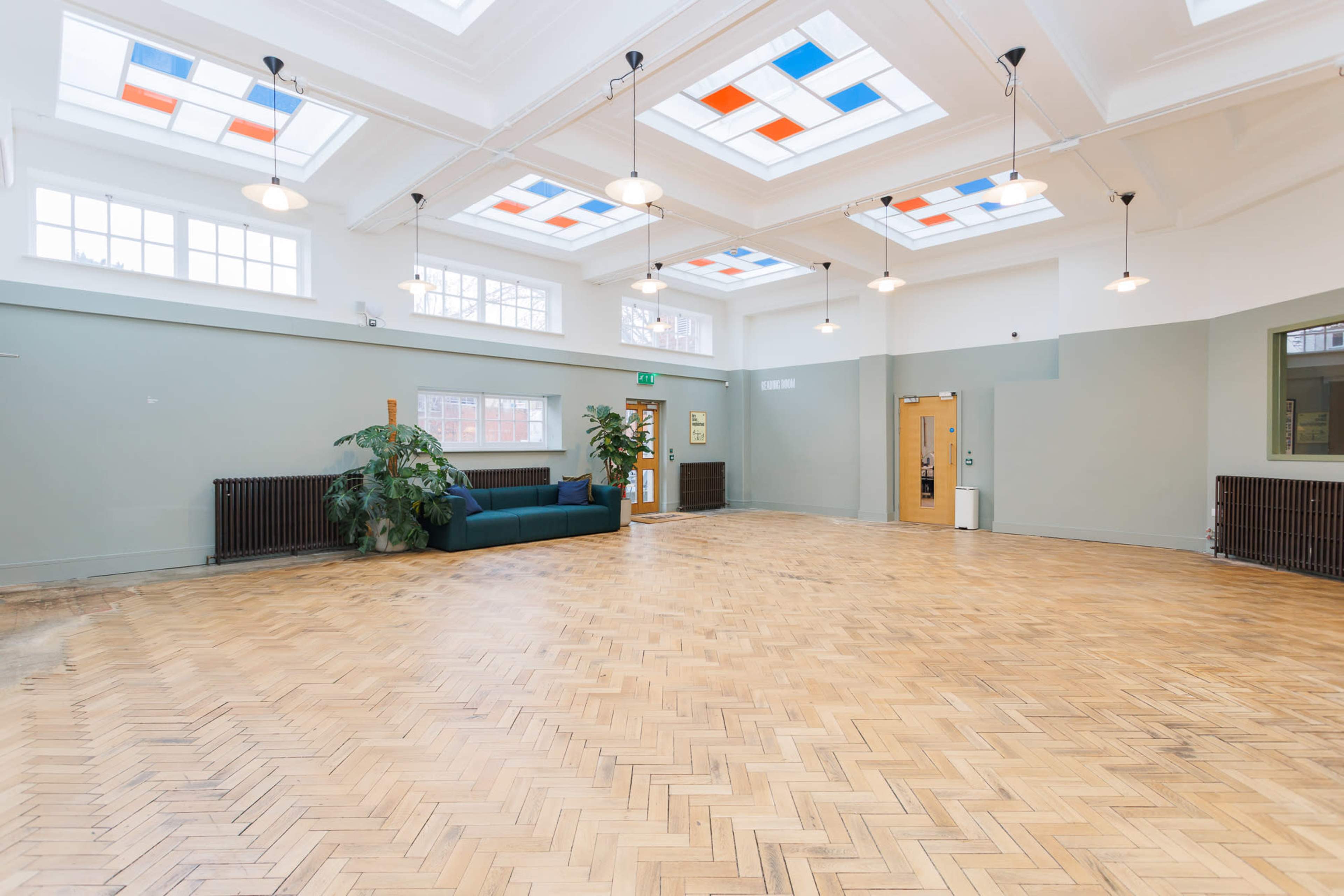 The image shows a spacious room with hardwood herringbone flooring, large windows featuring colored panes, and a green sofa against one wall.