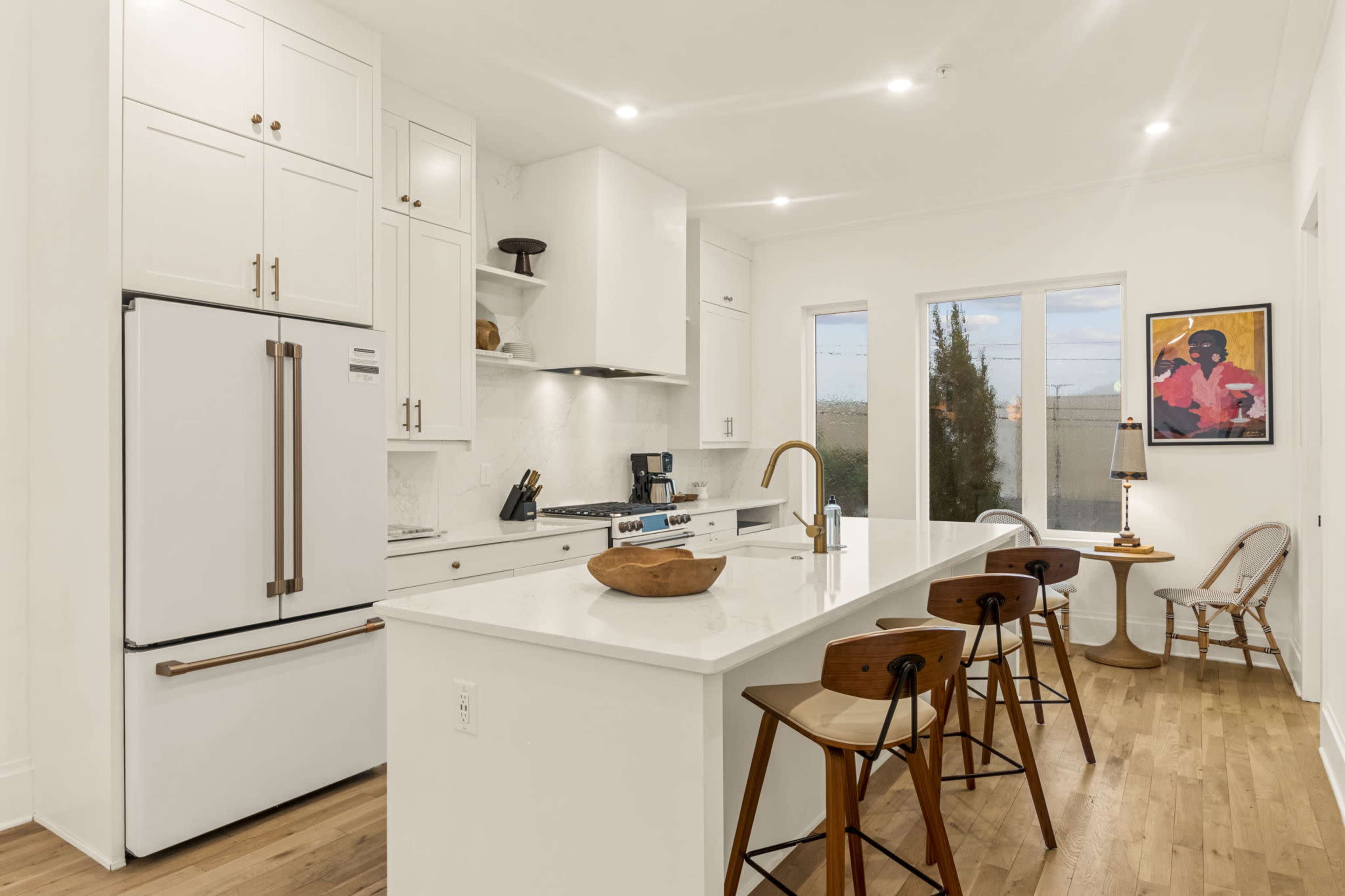 A modern kitchen features white cabinetry, a marble island with seating, and large windows allowing natural light.